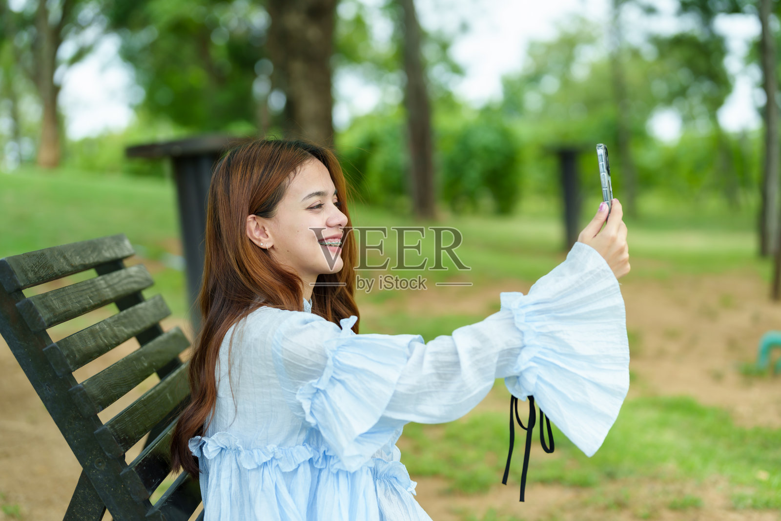 美丽的亚洲女性在度假，坐在绿色公园的木长椅上，微笑着，手持智能手机自拍，穿着时尚的浅蓝色连衣裙，享受户外休闲时光。照片摄影图片