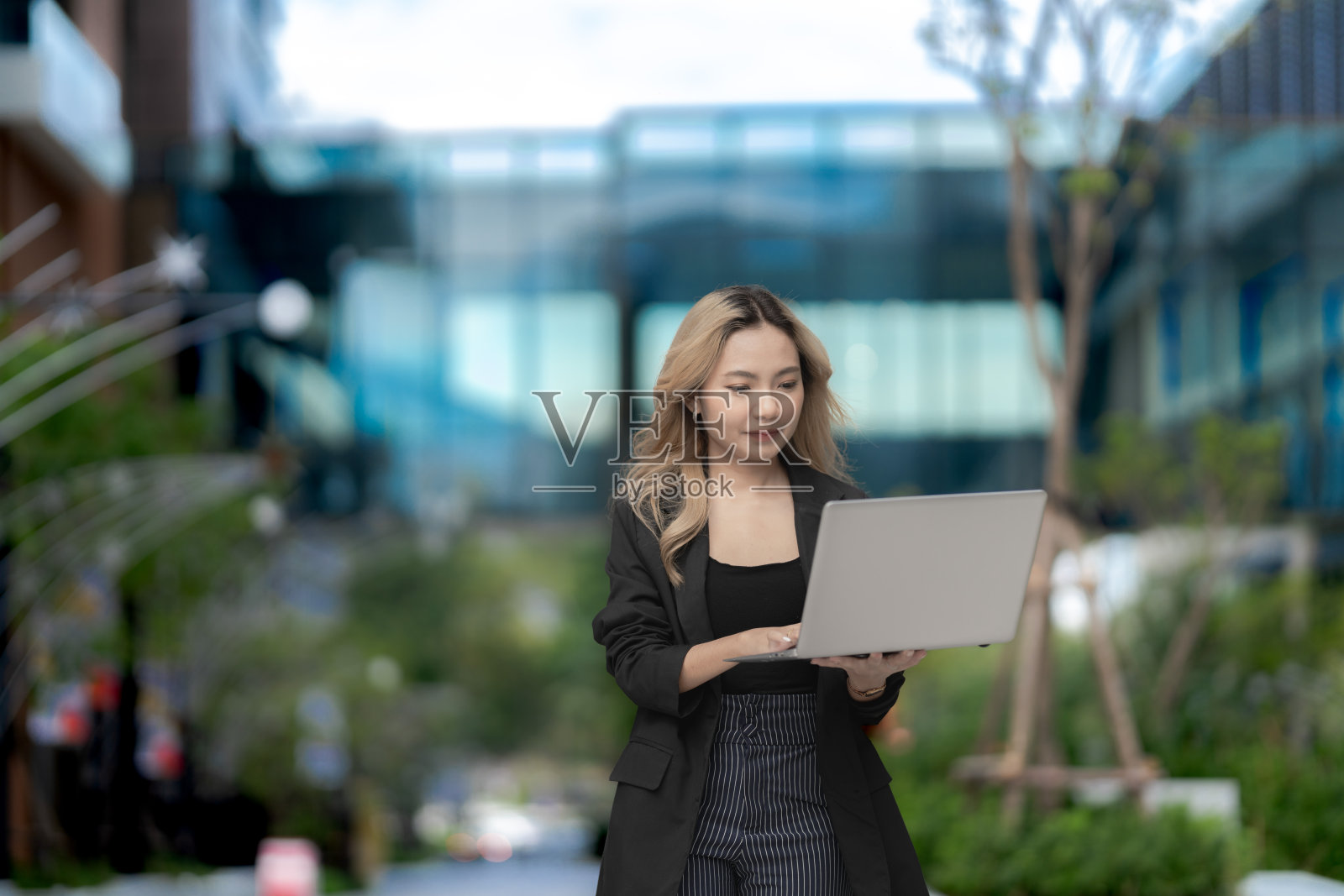 自信的女商人在户外走动，手持笔记本电脑，展示现代科技和职业抱负。城市绿化与创新的结合，体现了充满活力的生活方式。照片摄影图片