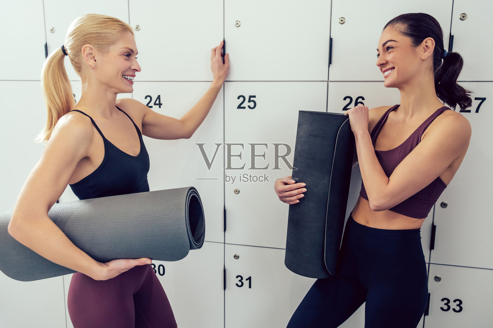 两位年轻女性在健身房的储物柜旁边拿着卷好的垫子照片摄影图片
