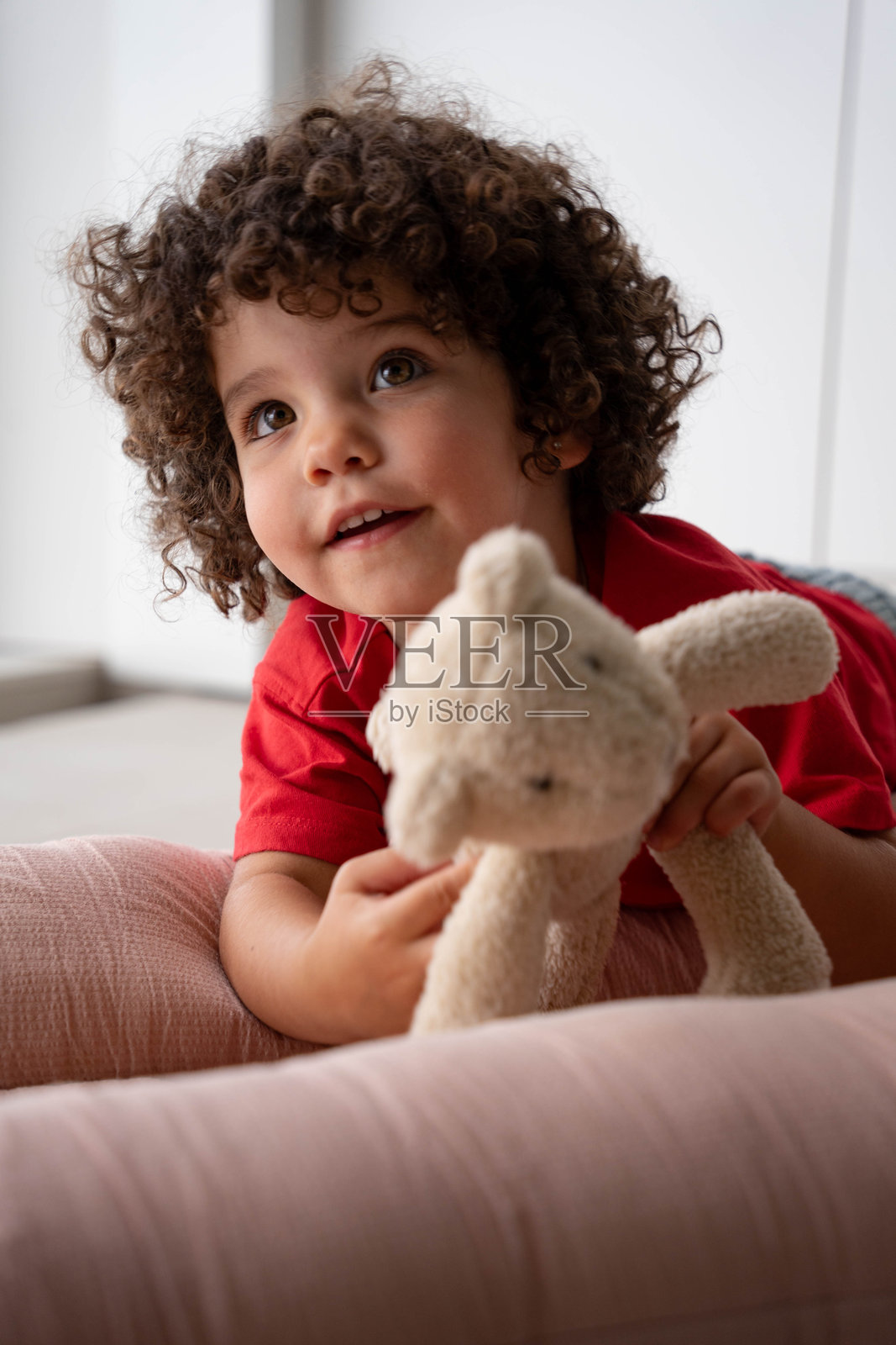 一个快乐的卷发孩子在窗边抱着一只泰迪熊，窗外是美丽的海洋景色。照片摄影图片