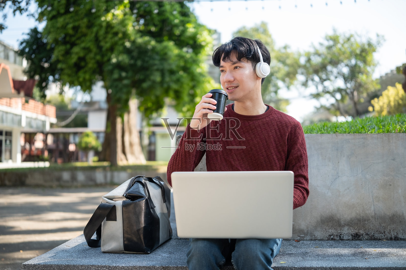 无忧无虑的亚洲男学生在校园公园里一边喝咖啡一边使用笔记本电脑。照片摄影图片