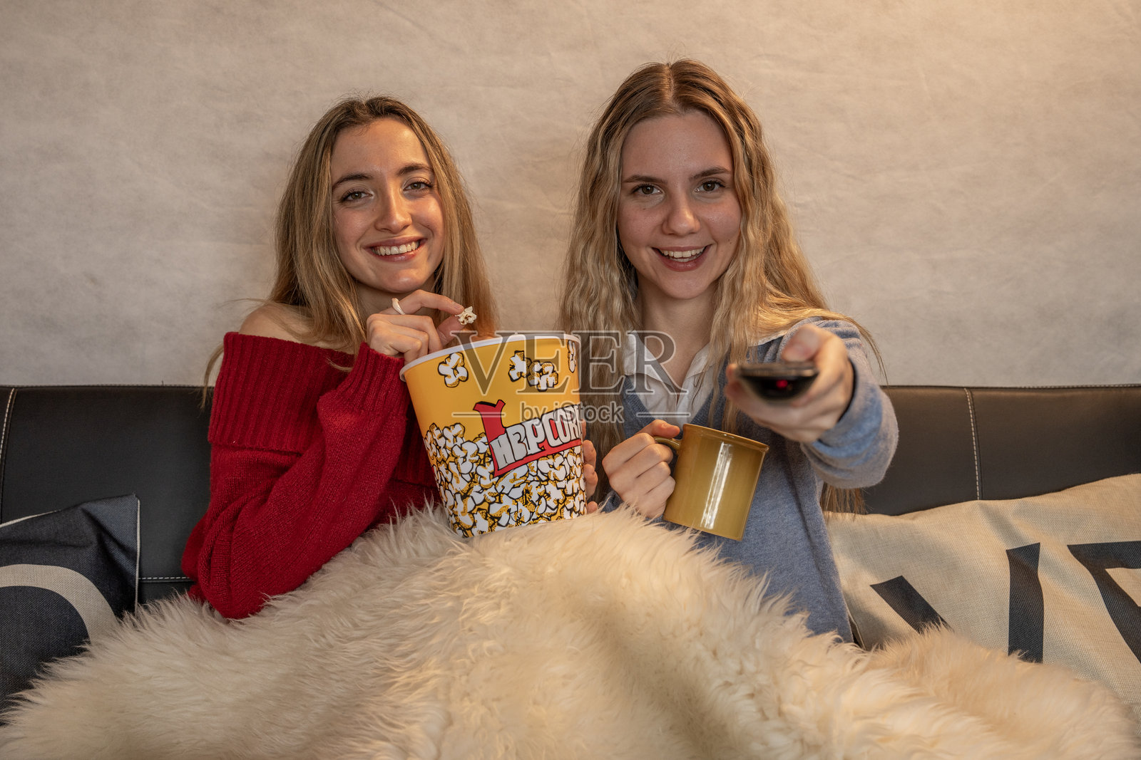 两个微笑的年轻女性在看电视，吃着爆米花，手里拿着遥控器。照片摄影图片