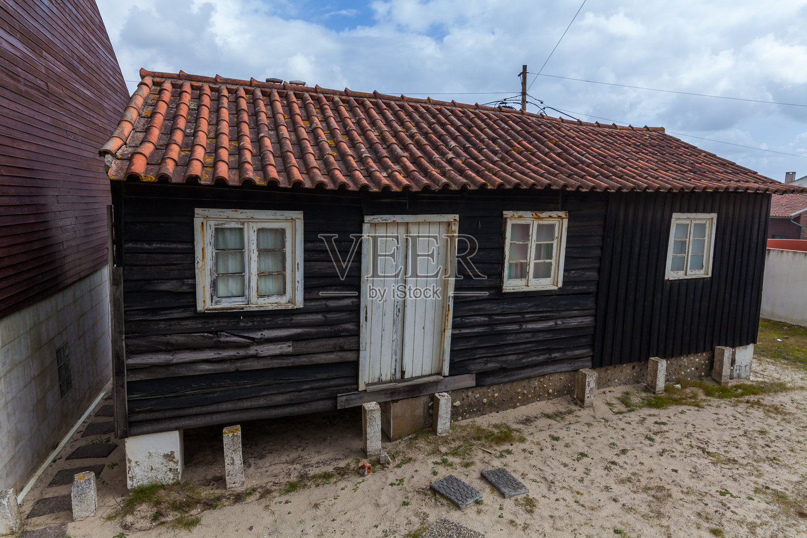 一座小巧、风化的木屋，红色瓦屋顶和白色边框，矗立在沙地上，背景是一片多云的天空。照片摄影图片