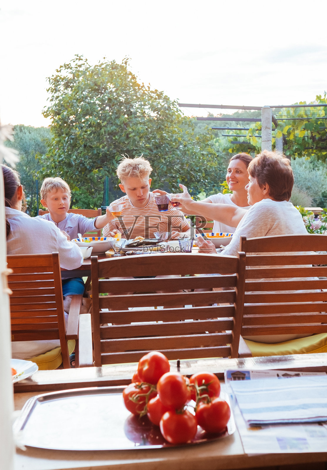家庭在阳光明媚的露台上享用户外餐，四周绿意盎然，分享欢笑与亲密时刻，前景中的新鲜番茄象征着农场到餐桌的生活方式和简单。照片摄影图片