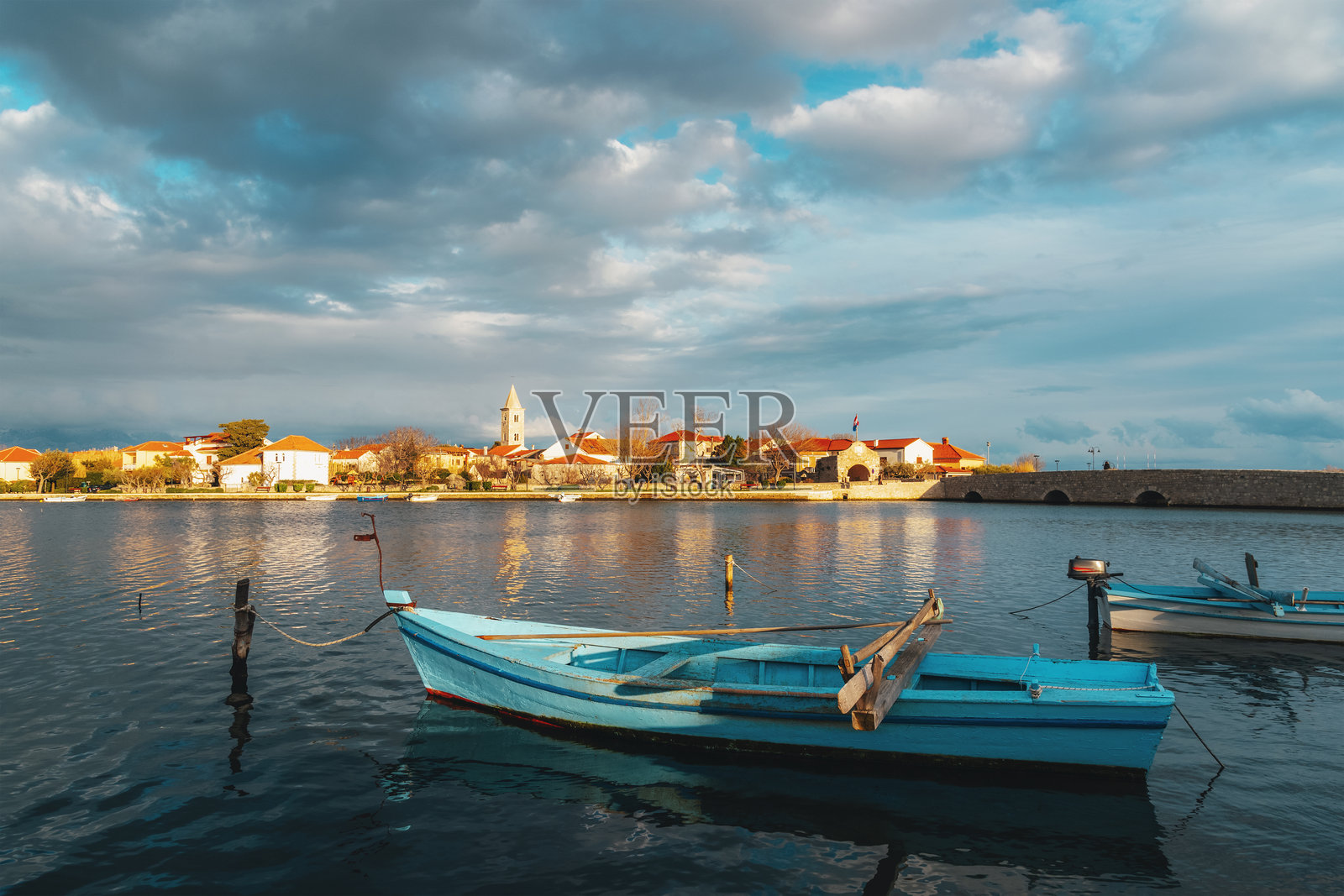 宁镇，克罗地亚，宁静的海岸风光，蓝色木制渔船在平静的水面上，历史悠久的村庄和教堂塔楼在日落时分映衬着戏剧性的云彩。照片摄影图片
