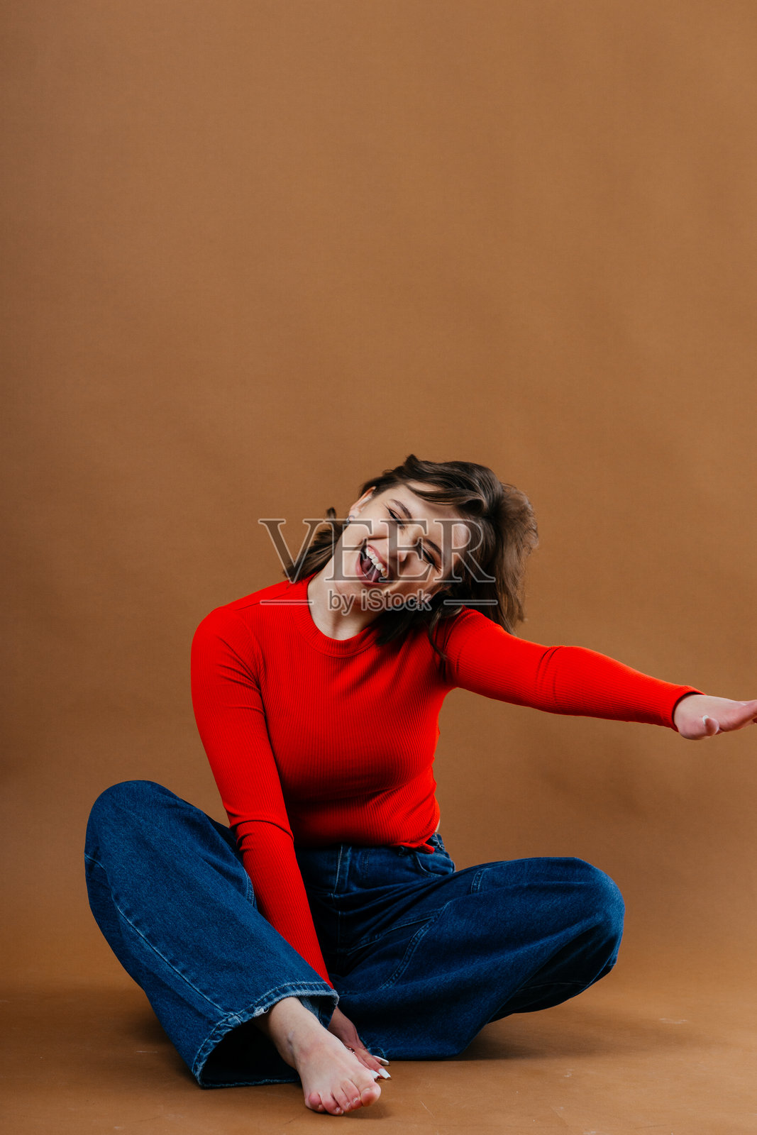 一位时尚的年轻女性穿着大胆的红色上衣和宽腿牛仔裤，在温暖的棕色背景前摆出动感而艺术的姿势。照片摄影图片