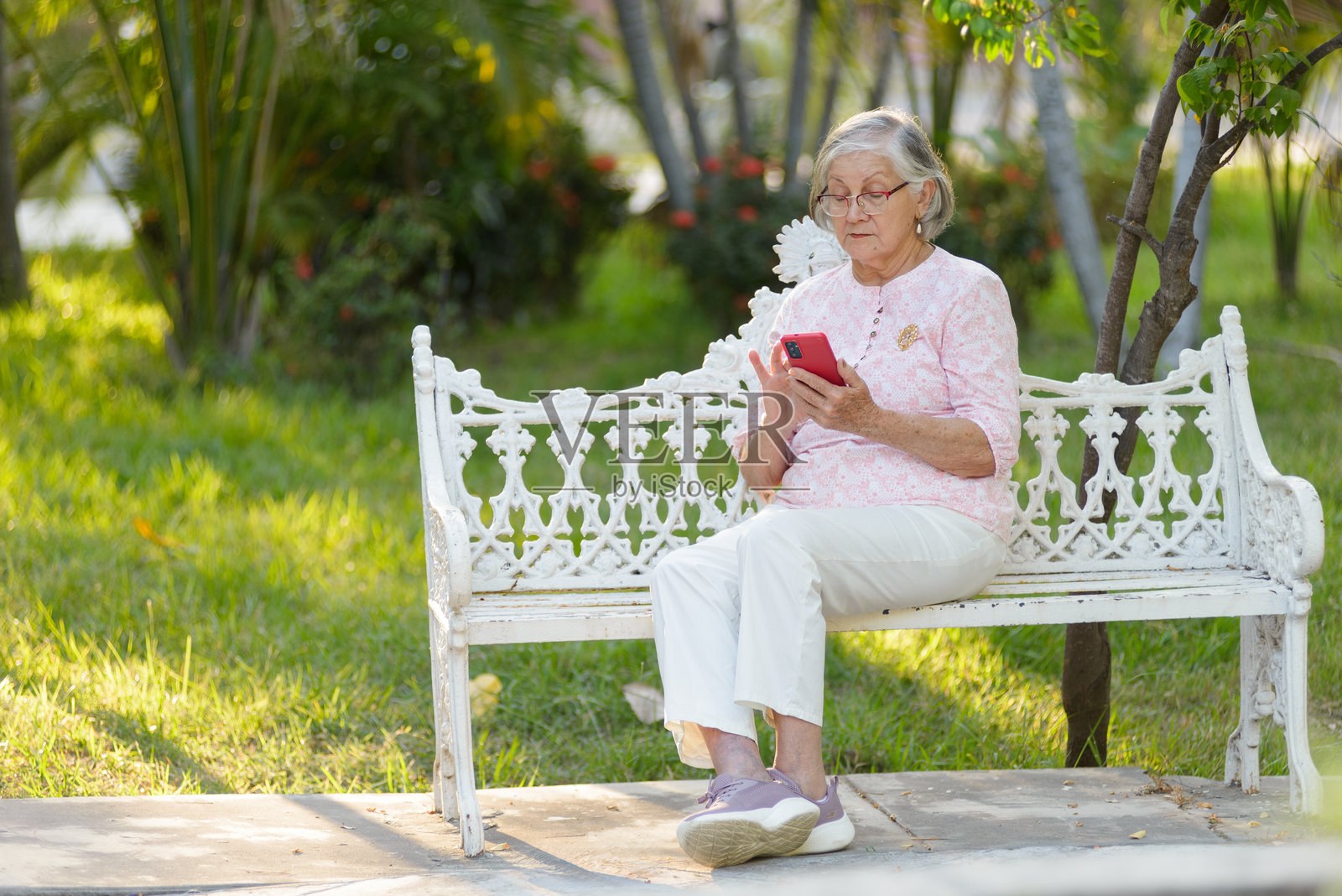 一位戴着眼镜的灰发老妇人坐在公园长椅上，专注于她的智能手机。照片摄影图片