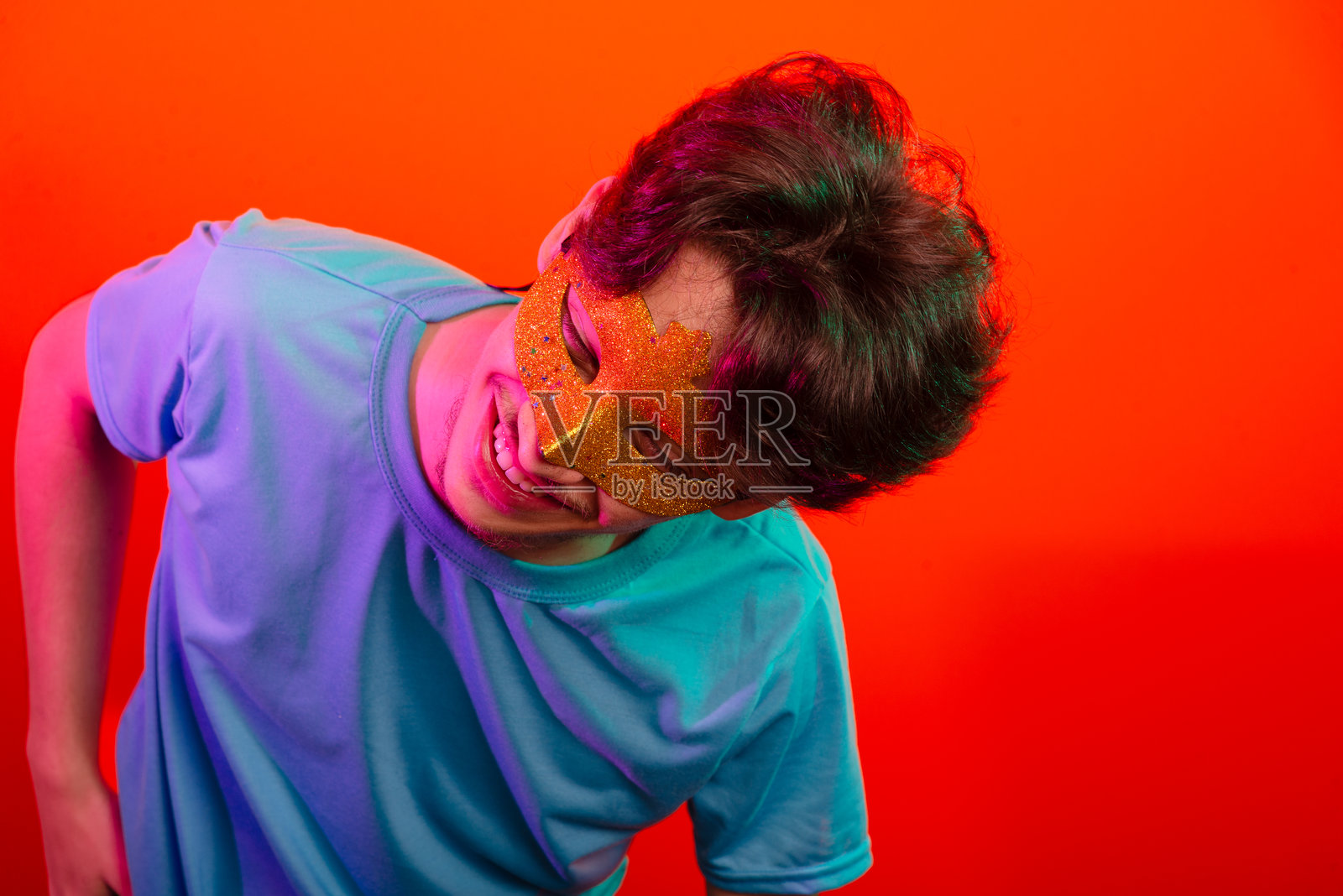 快乐有趣的年轻人戴着狂欢节面具，朝着镜头看并做手势。背景为霓虹橙色。照片摄影图片