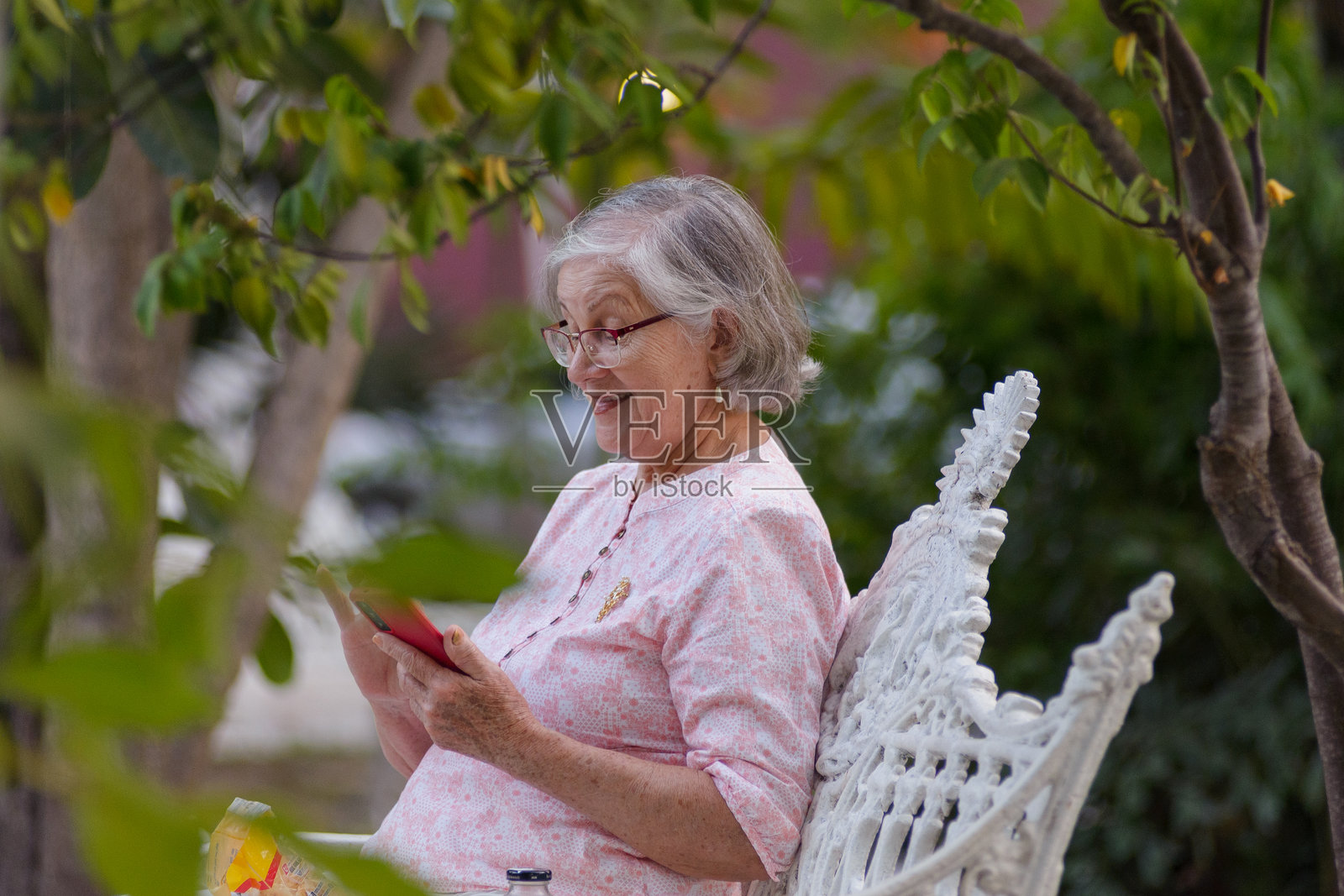 一位戴着眼镜的灰发老妇人坐在公园长椅上，专注于她的智能手机。照片摄影图片