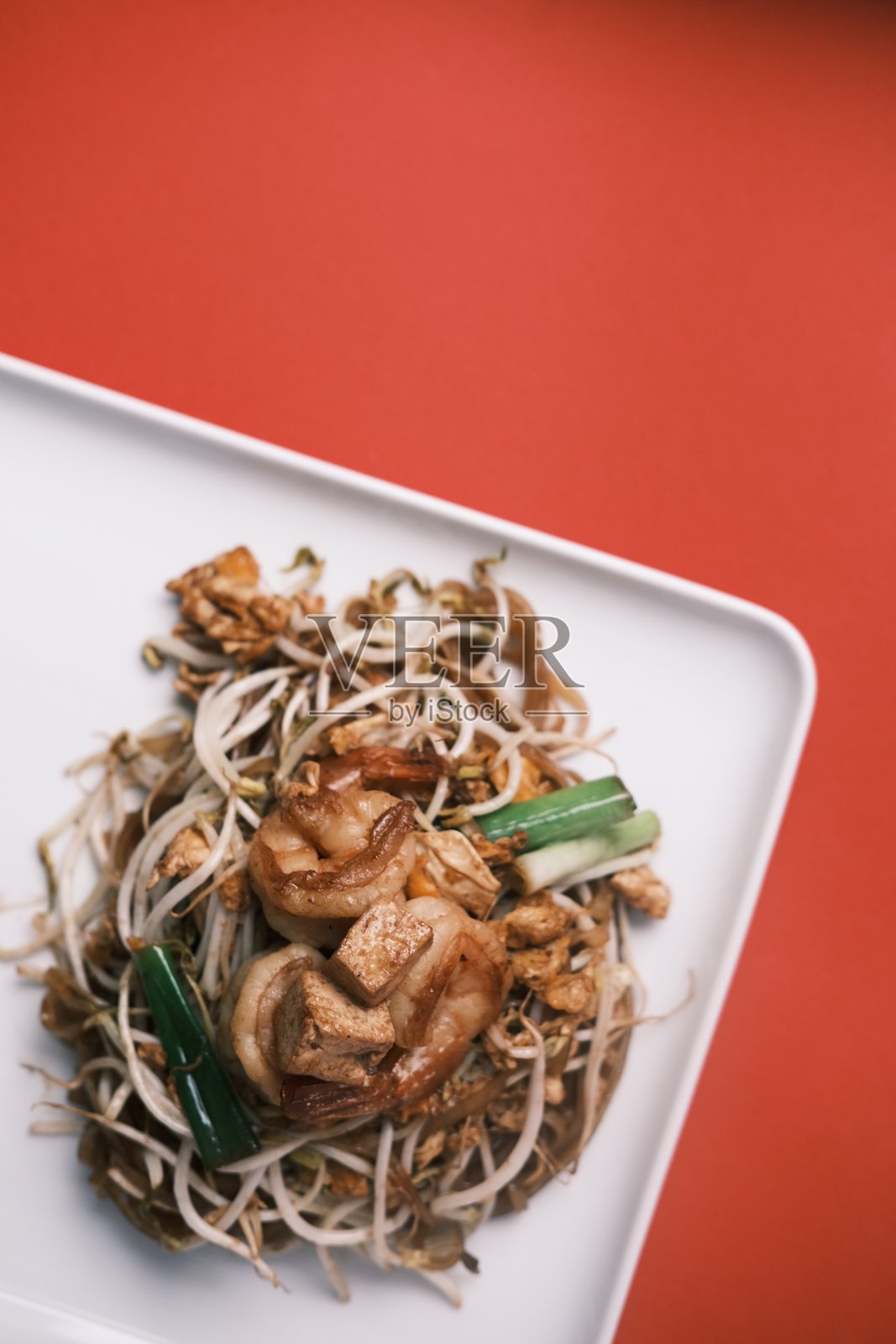 特写传统泰式虾仁炒米粉，配有豆芽和青葱，摆放在白色长方形盘子上，背景为鲜艳的红色，展现正宗泰国街头美食和亚洲料理概念。照片摄影图片