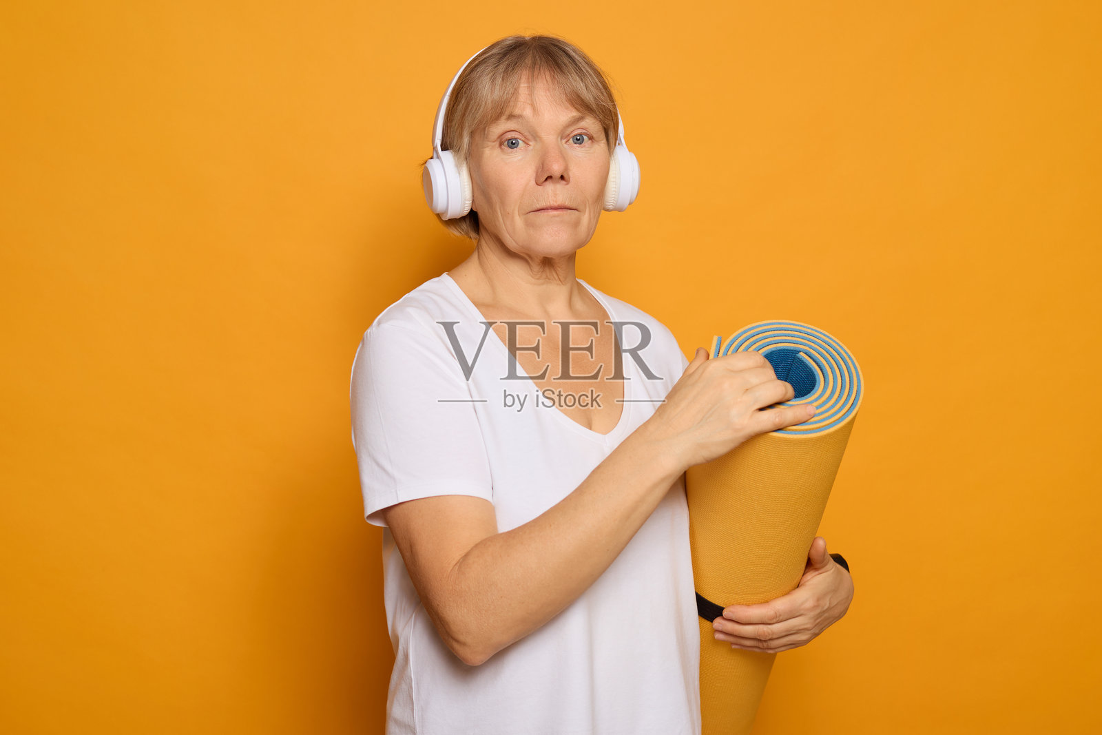 一位穿着休闲服的女性站在鲜艳的黄色背景前，手里拿着卷好的瑜伽垫，戴着耳机，准备开始她的锻炼。照片摄影图片