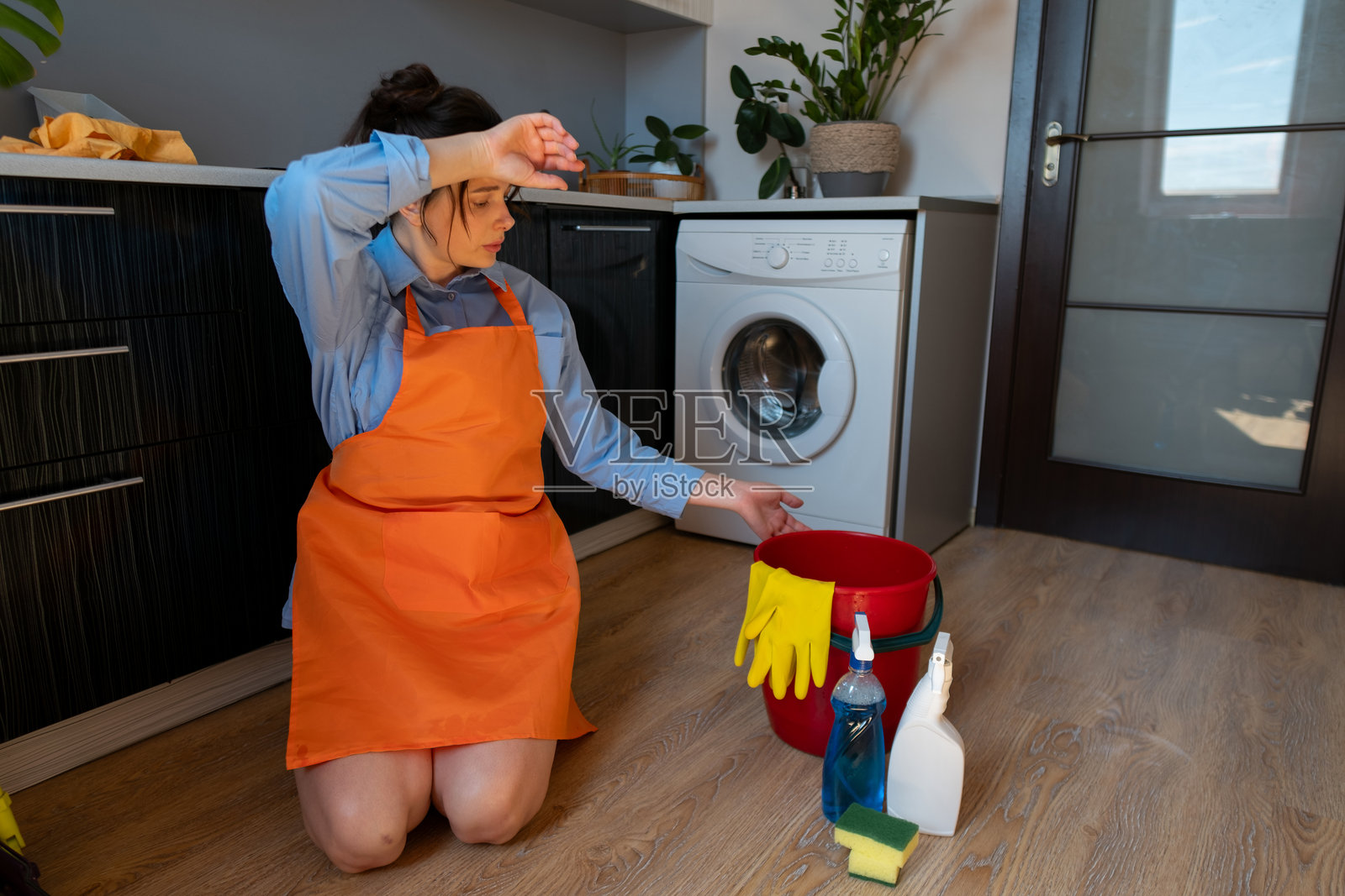 疲惫的女人坐在地板上，捂着头。定期清洁地板可以防止顽固污渍，并使未来的清洁工作更轻松。照片摄影图片