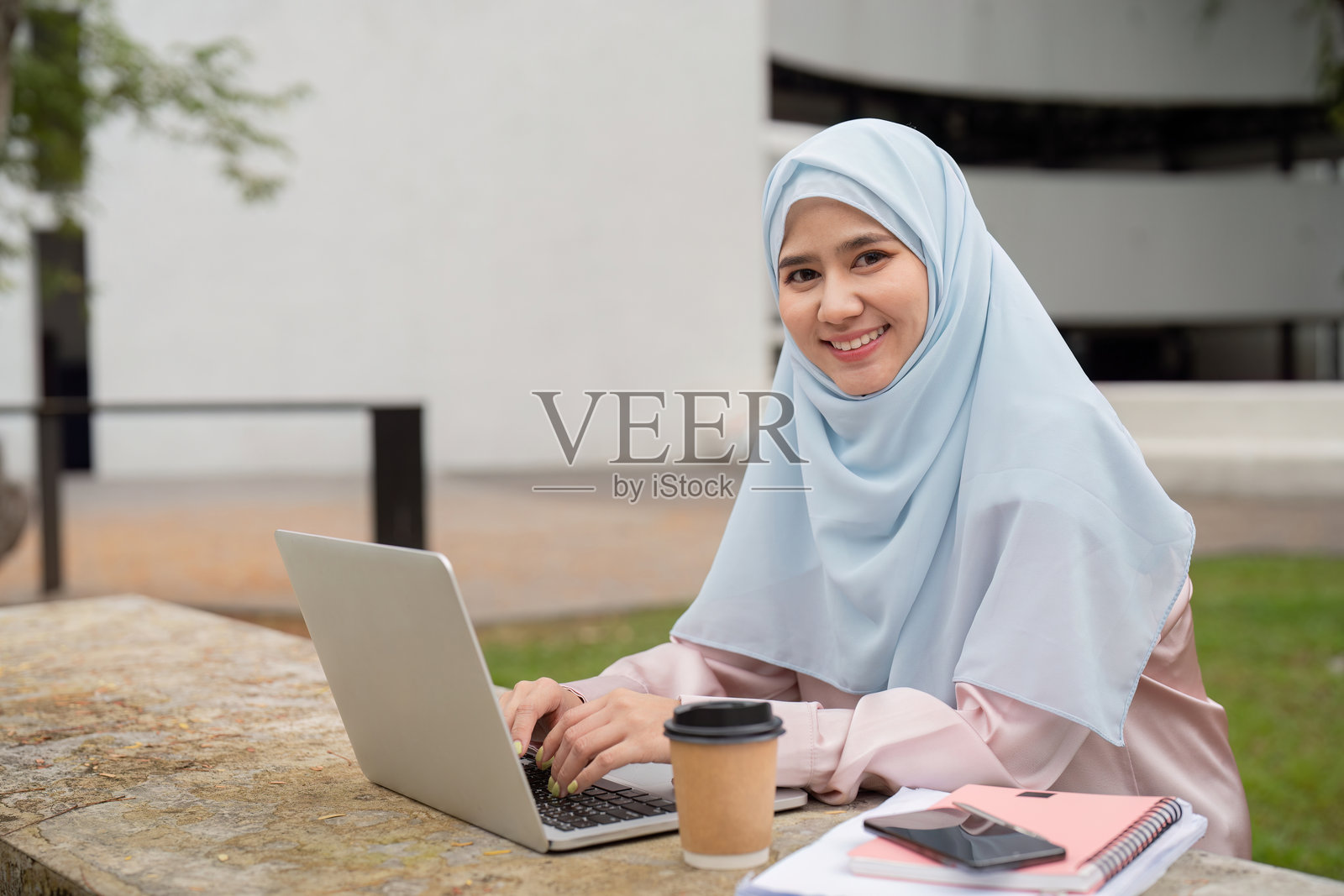 微笑的穆斯林女商人在户外戴着头巾，喝着咖啡，使用笔记本电脑。照片摄影图片