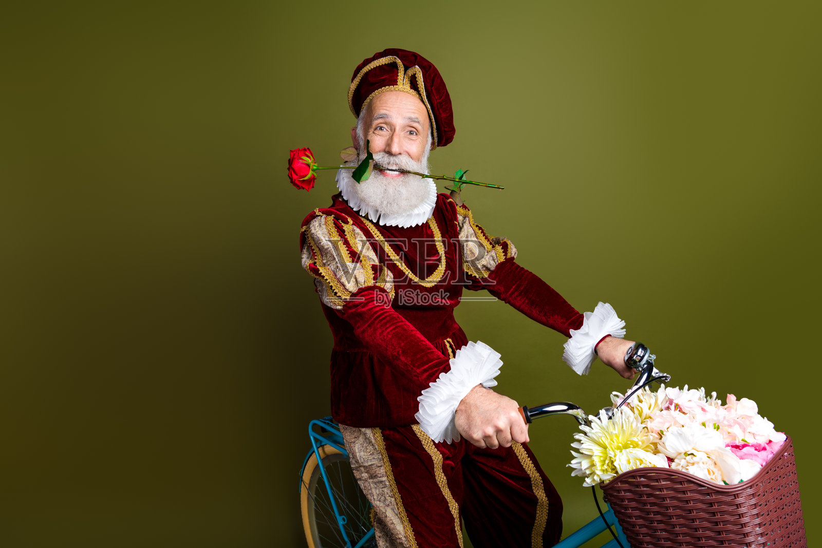 身穿中世纪服装的成熟男子骑着自行车，手持红玫瑰，篮子里装满鲜花，背景为卡其色。照片摄影图片