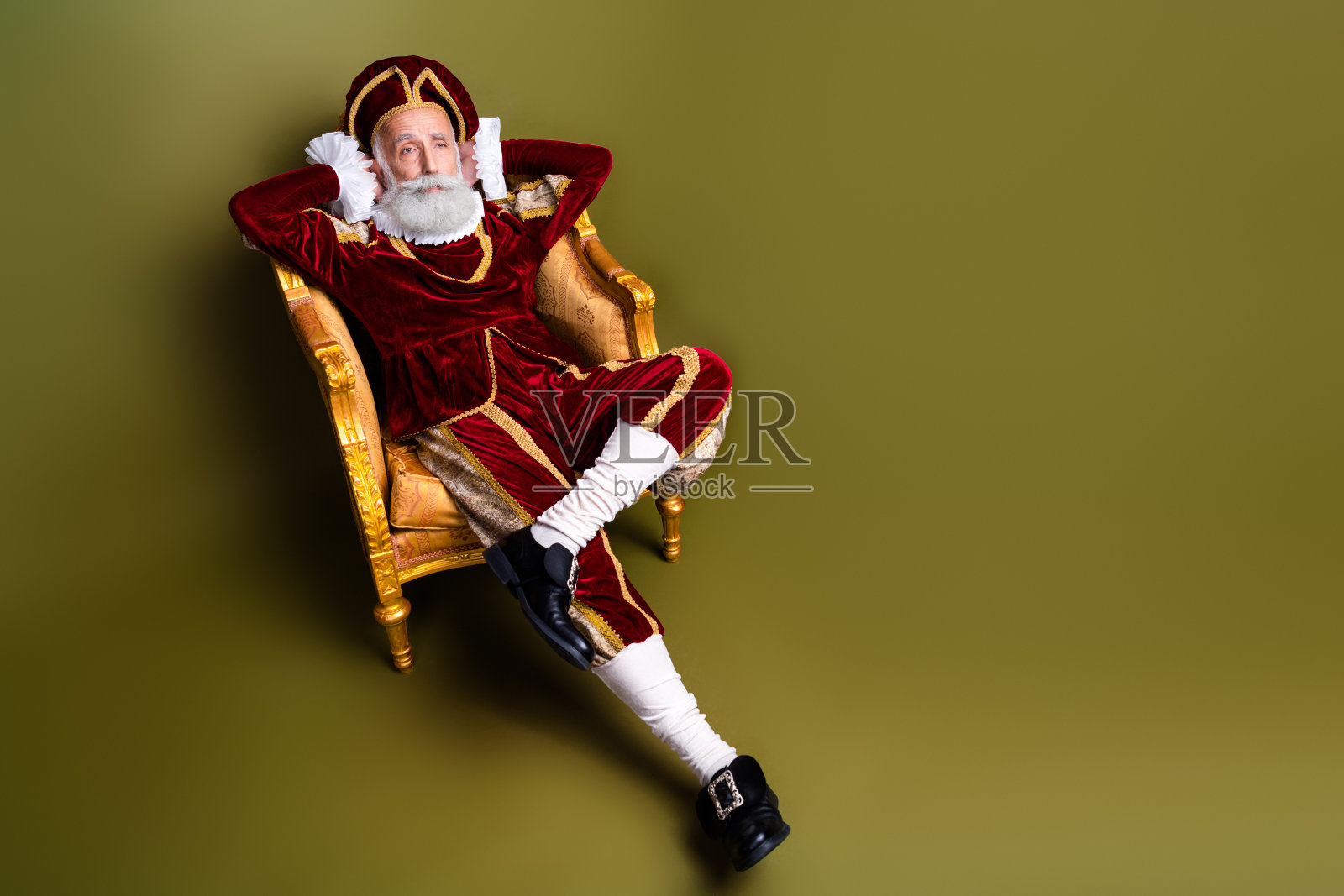 穿着皇家红色服装的老年男子懒散地坐在古董椅子上，背景为卡其色。照片摄影图片
