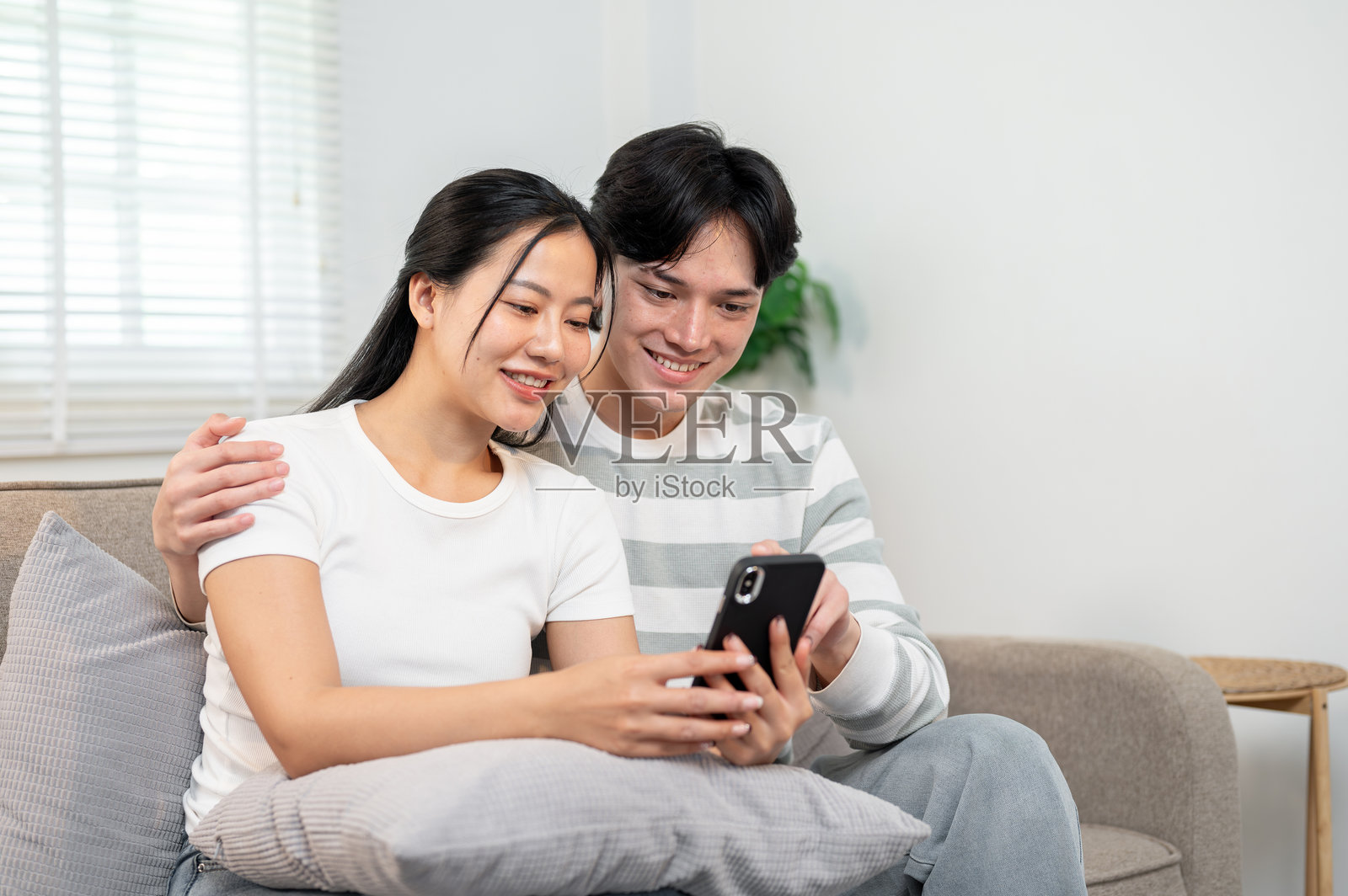 一对快乐的亚洲夫妇坐在客厅的沙发上，一起观看手机上的视频。照片摄影图片