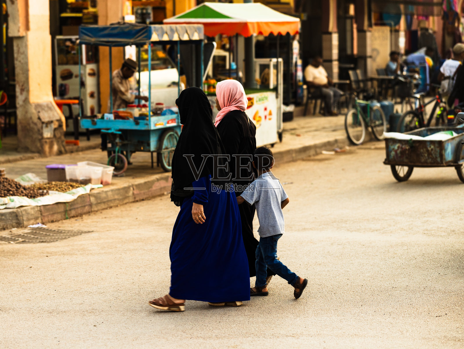 两位身着传统服饰的女性在日落时分带着一个孩子走过街市。照片摄影图片