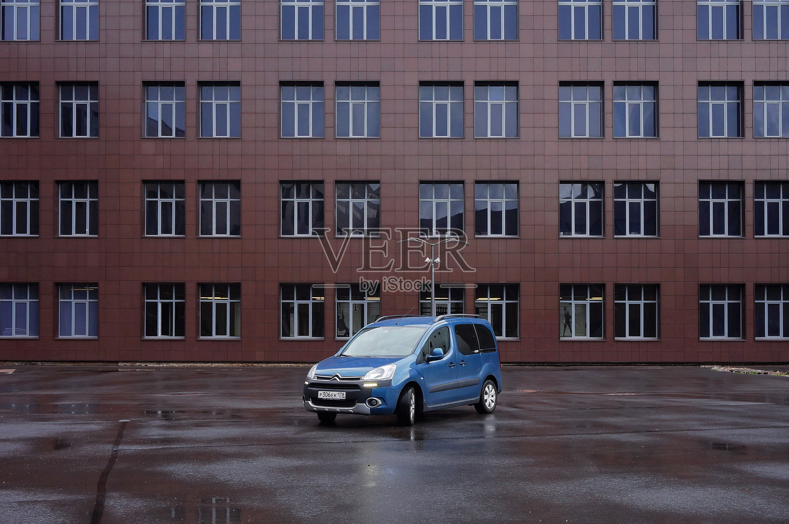 蓝色雪铁龙Berlingo B9在雨天停放。三分之四前视图。背景是一座大型棕色办公楼,具有重复的窗户图案。照片摄影图片
