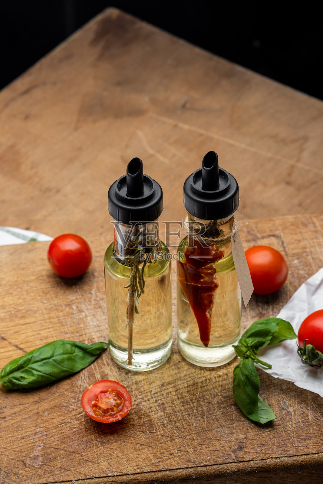 透明玻璃瓶装的橄榄油，里面有红辣椒、黑胡椒粒和迷迭香，背景是木质纹理，旁边有罗勒叶、樱桃番茄、意大利调味酱和香料。照片摄影图片