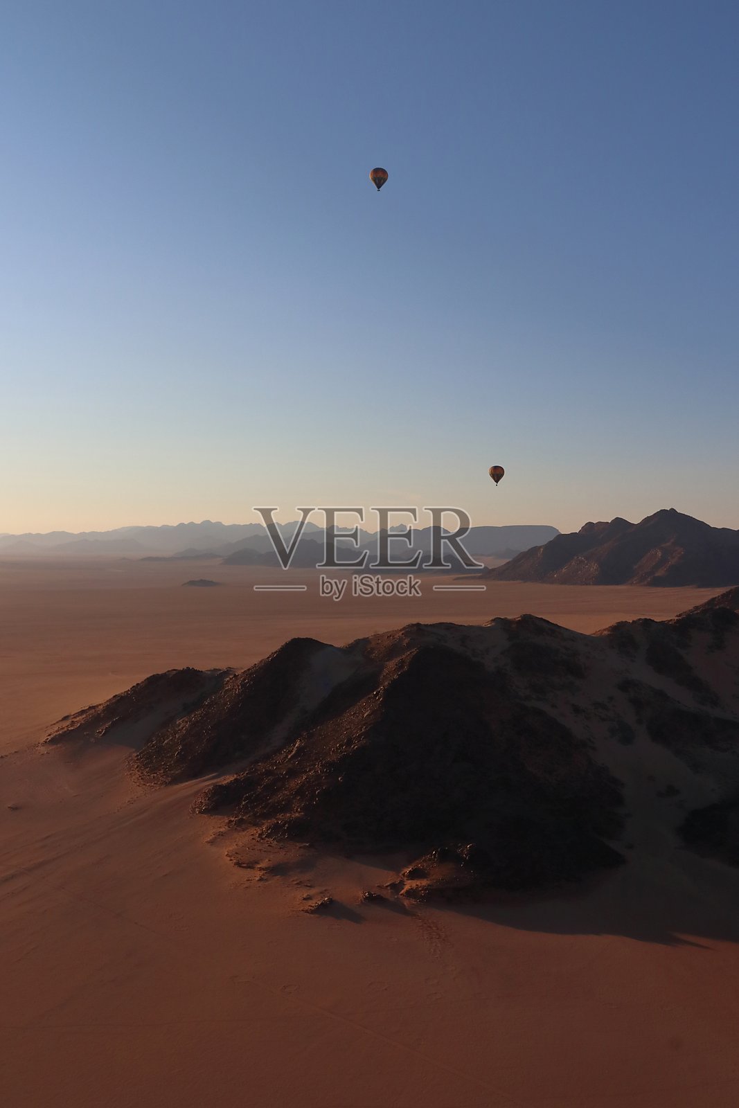 热气球在纳米比亚的纳米布沙漠上空漂浮，日出时分提供了对崎岖地貌、金色沙丘和远处山脊的空中视角，沐浴在柔和的晨光中。照片摄影图片