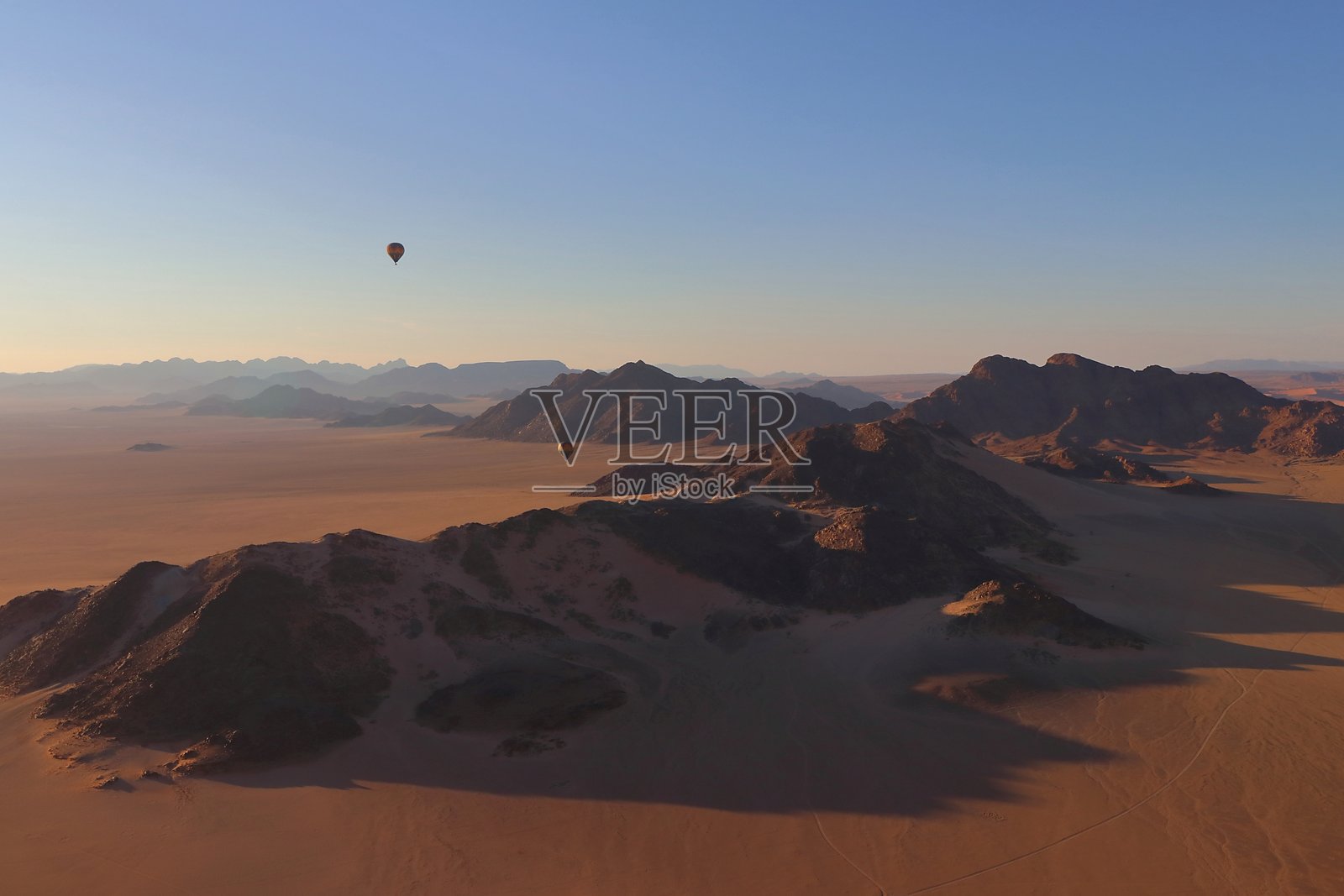 热气球在纳米比亚的纳米布沙漠上空漂浮，日出时分提供了对崎岖地貌、金色沙丘和远处山脊的空中视角，沐浴在柔和的晨光中。照片摄影图片
