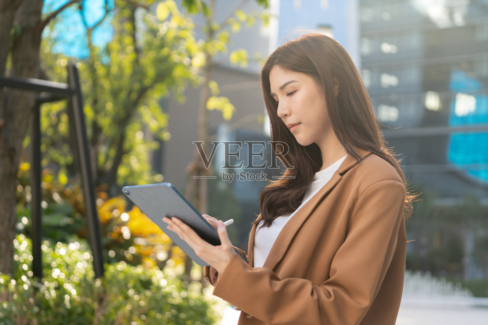 女性在现代城市环境中户外使用平板电脑，周围环绕着绿植和建筑，展示了科技与自然的融合。照片摄影图片