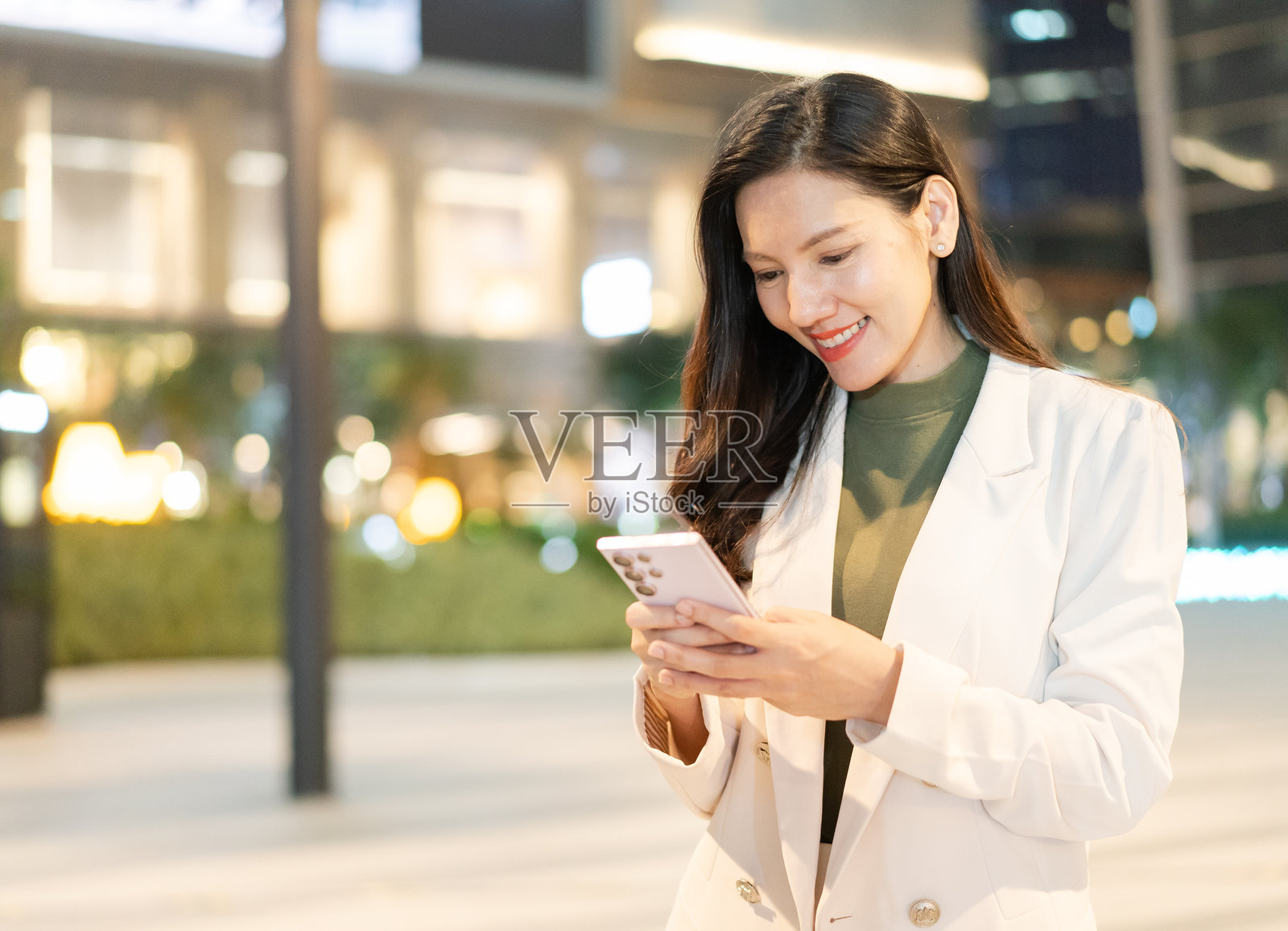 穿着白色西装的女性在夜晚户外微笑着使用智能手机，背景是城市灯光，营造出充满活力的氛围。照片摄影图片