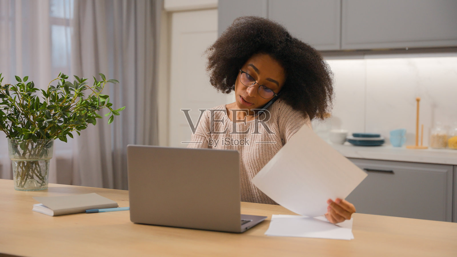 非裔美国女性多任务处理的商业女性，自由职业者，企业家，在家厨房远程工作，使用笔记本电脑，女性族裔女孩与手机通话，咨询客户，搜索数据，处理文书工作。照片摄影图片