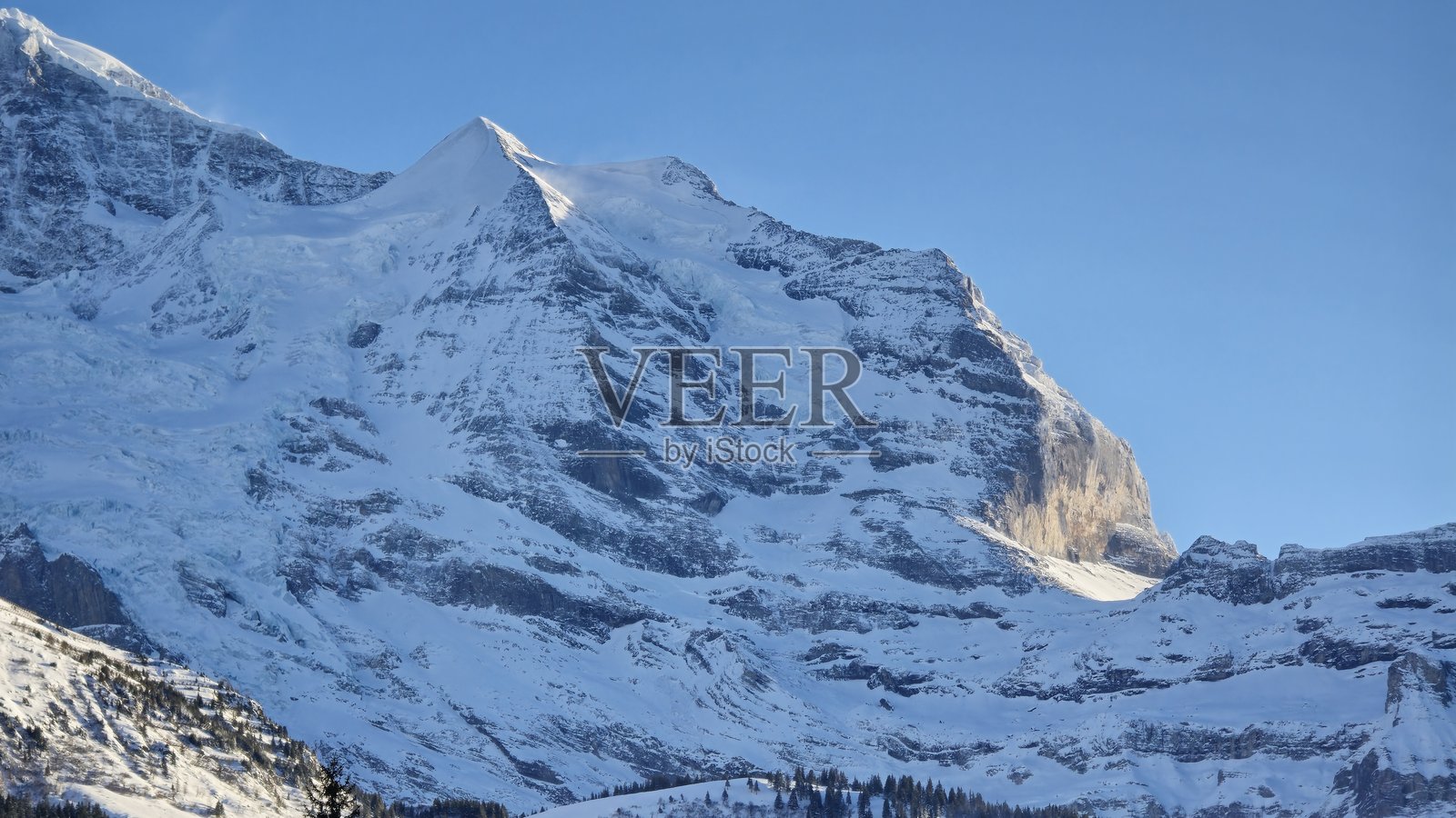 一座雄伟的雪山峰在晴朗的蓝天映衬下拔地而起。阳光照耀着崎岖的悬崖和冰冷的坡面，营造出令人叹为观止的高山风光，散发着清新的冬季气息。照片摄影图片