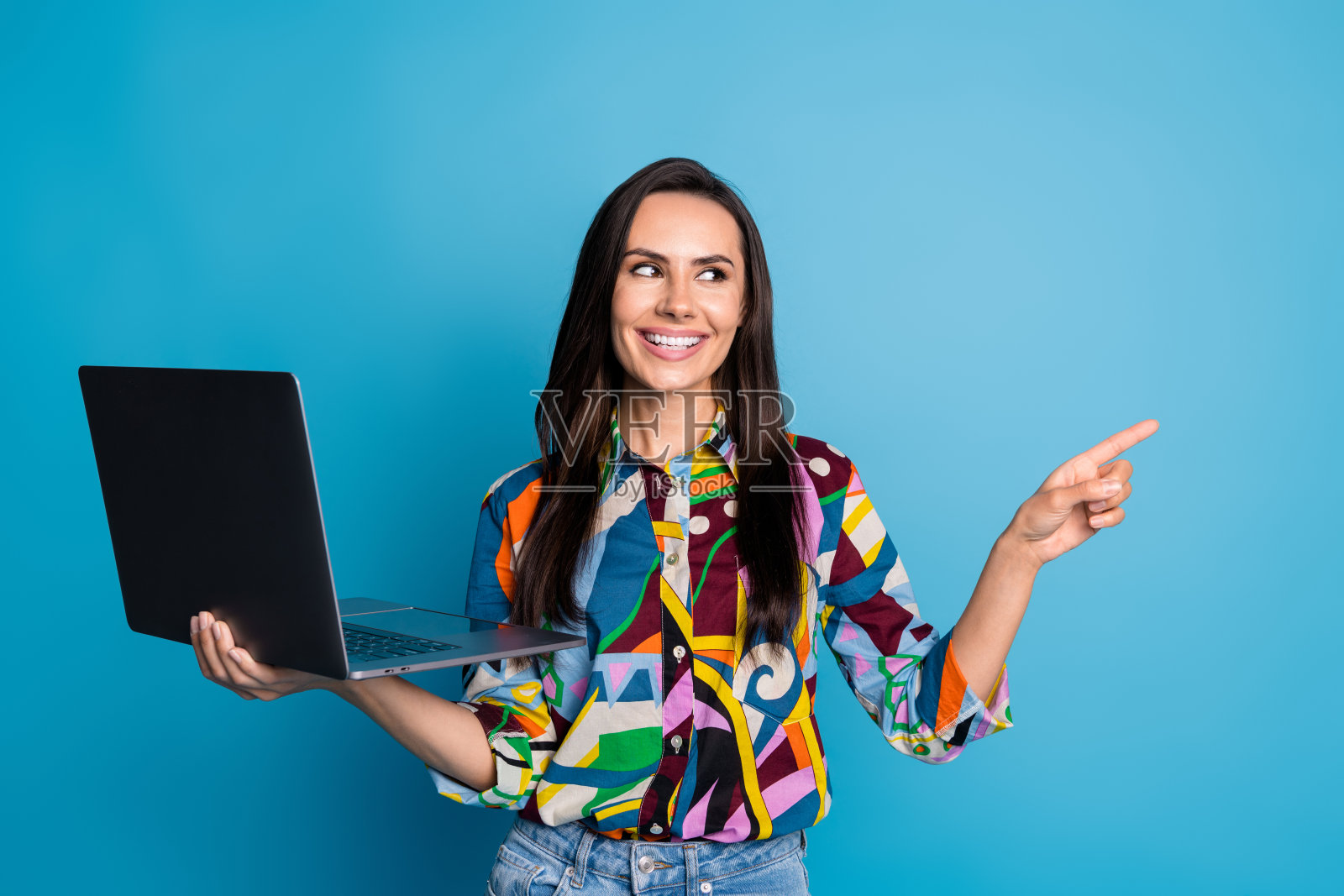 可爱的女性，直发，穿着印花衬衫，手持笔记本电脑，目光注视空旷的空间，背景为蓝色。照片摄影图片