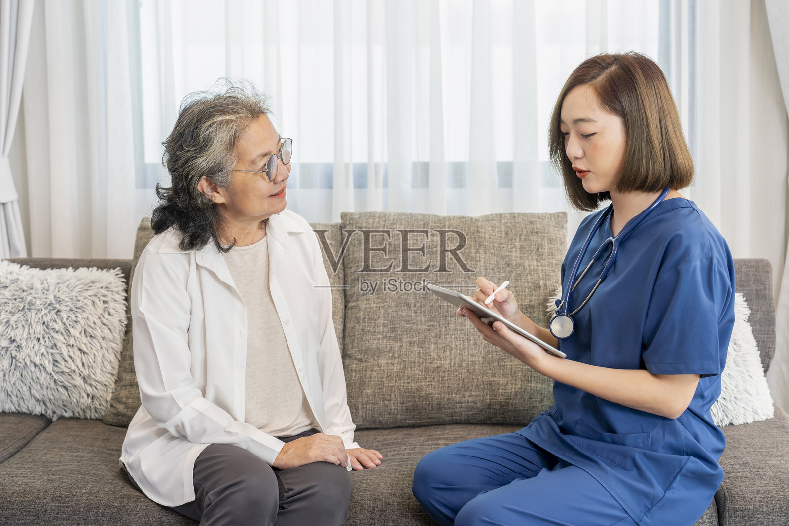 穿着制服的亚洲女性护士在家中探访一位老年患者，使用平板电脑进行评估、收集数据并提供建议，老年健康护理，居家护理照片摄影图片