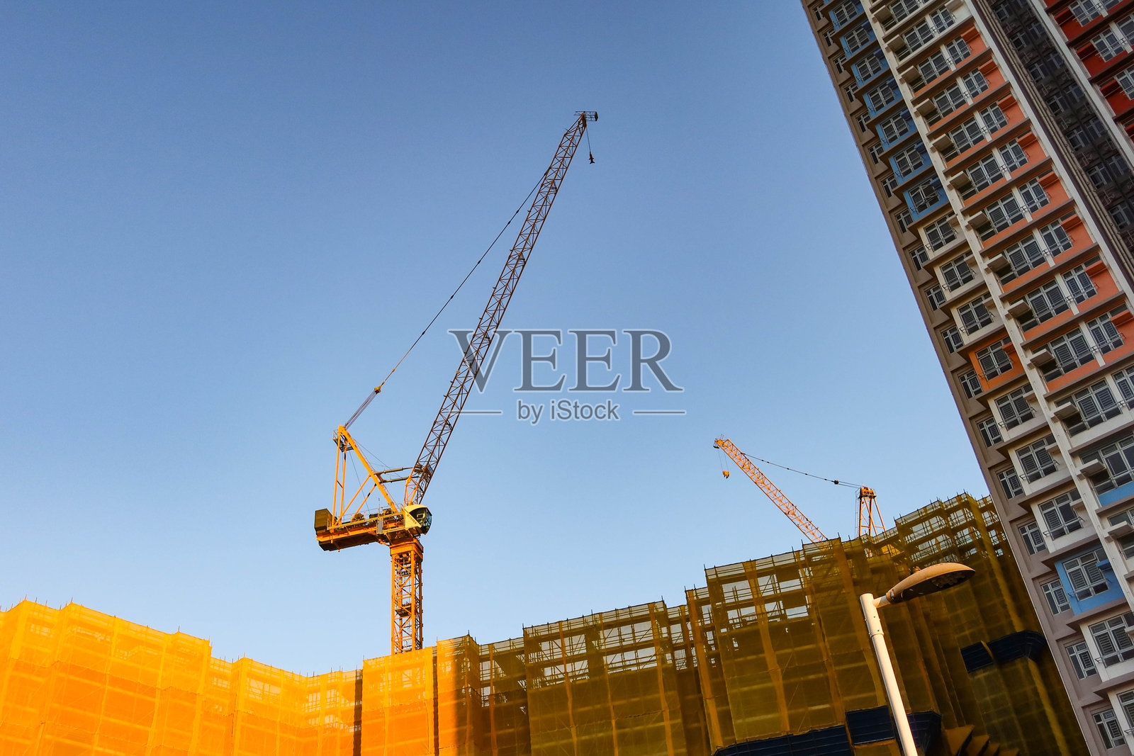 傍晚时分，蓝天映衬下，正在建设中的住宅楼群特写，塔吊和黄色安全网清晰可见。照片摄影图片