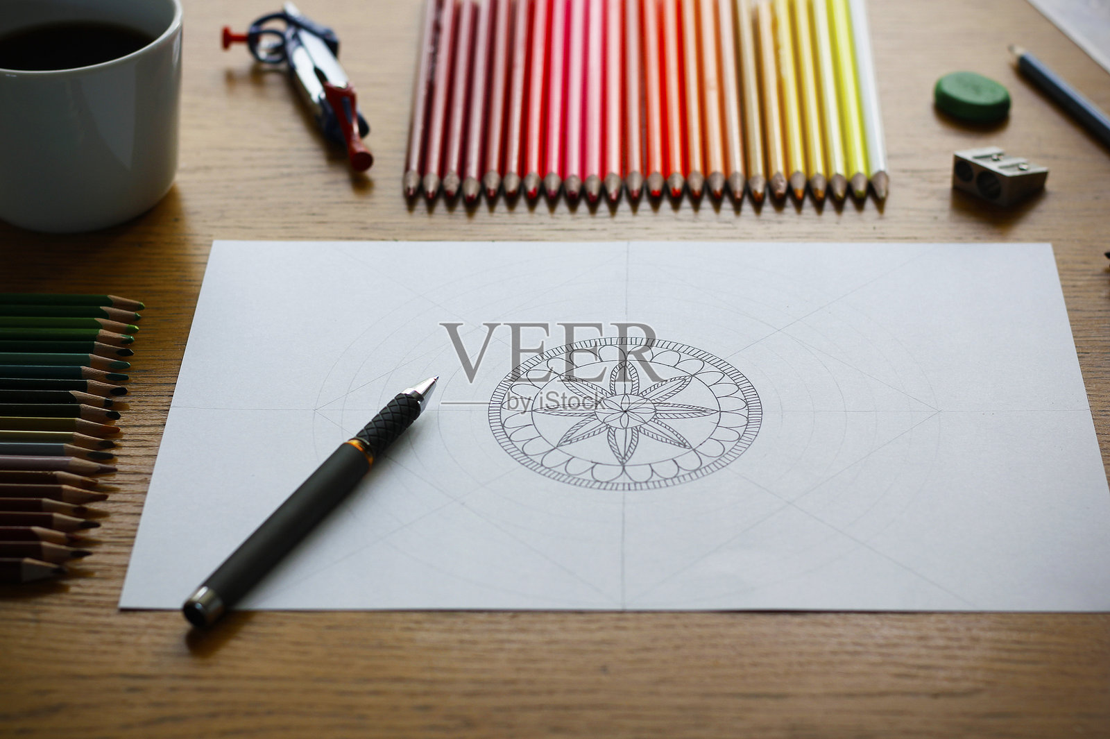 一幅绘制的曼荼罗图案在白纸上，放在木桌上，旁边有绘画工具和一杯咖啡。照片摄影图片