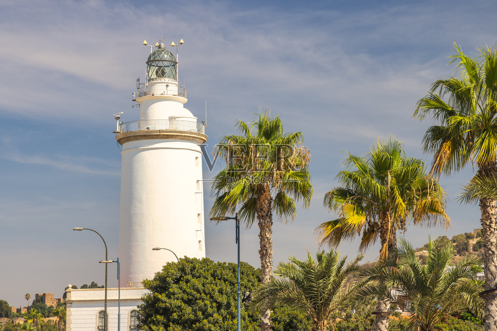 马拉加，西班牙安达卢西亚的海滨城市。白色灯塔在明亮的蓝天下高高耸立，周围环绕着郁郁葱葱的棕榈树。一个如画的海岸风景。照片摄影图片