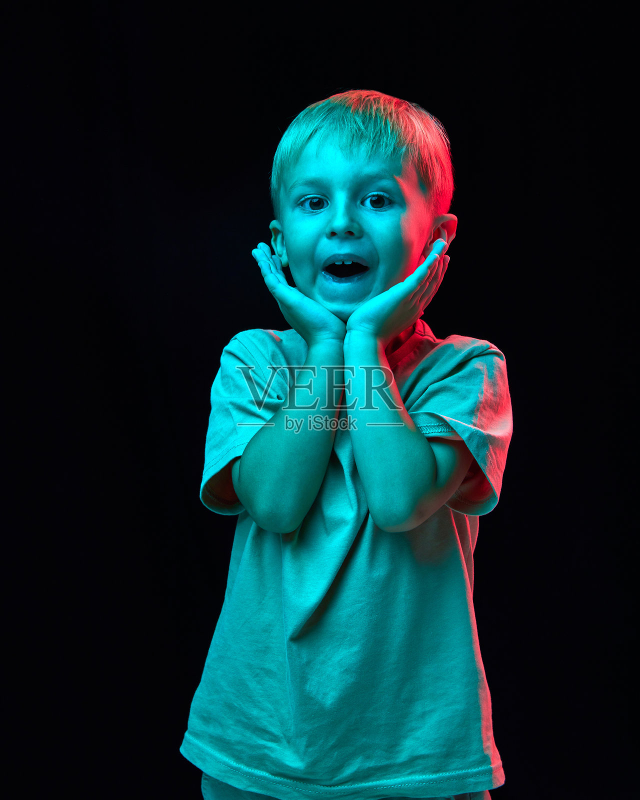 可爱小男孩的肖像，穿着休闲白色T恤，双手托着脸颊，微笑着，背景是黑色摄影棚中的霓虹灯光。照片摄影图片