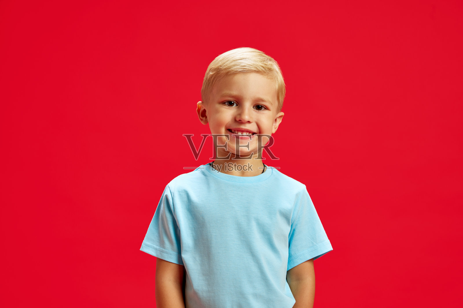 四岁小男孩的肖像，金色头发，面带愉快的微笑，穿着蓝色T恤，站在红色摄影背景前摆姿势。照片摄影图片