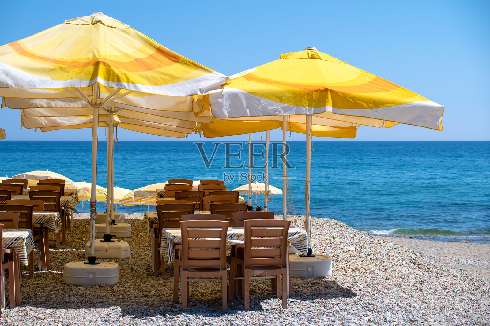 放松的海滨咖啡馆场景。木制桌椅在鲜艳的黄色伞下，俯瞰着迷人的碧蓝海洋。照片摄影图片