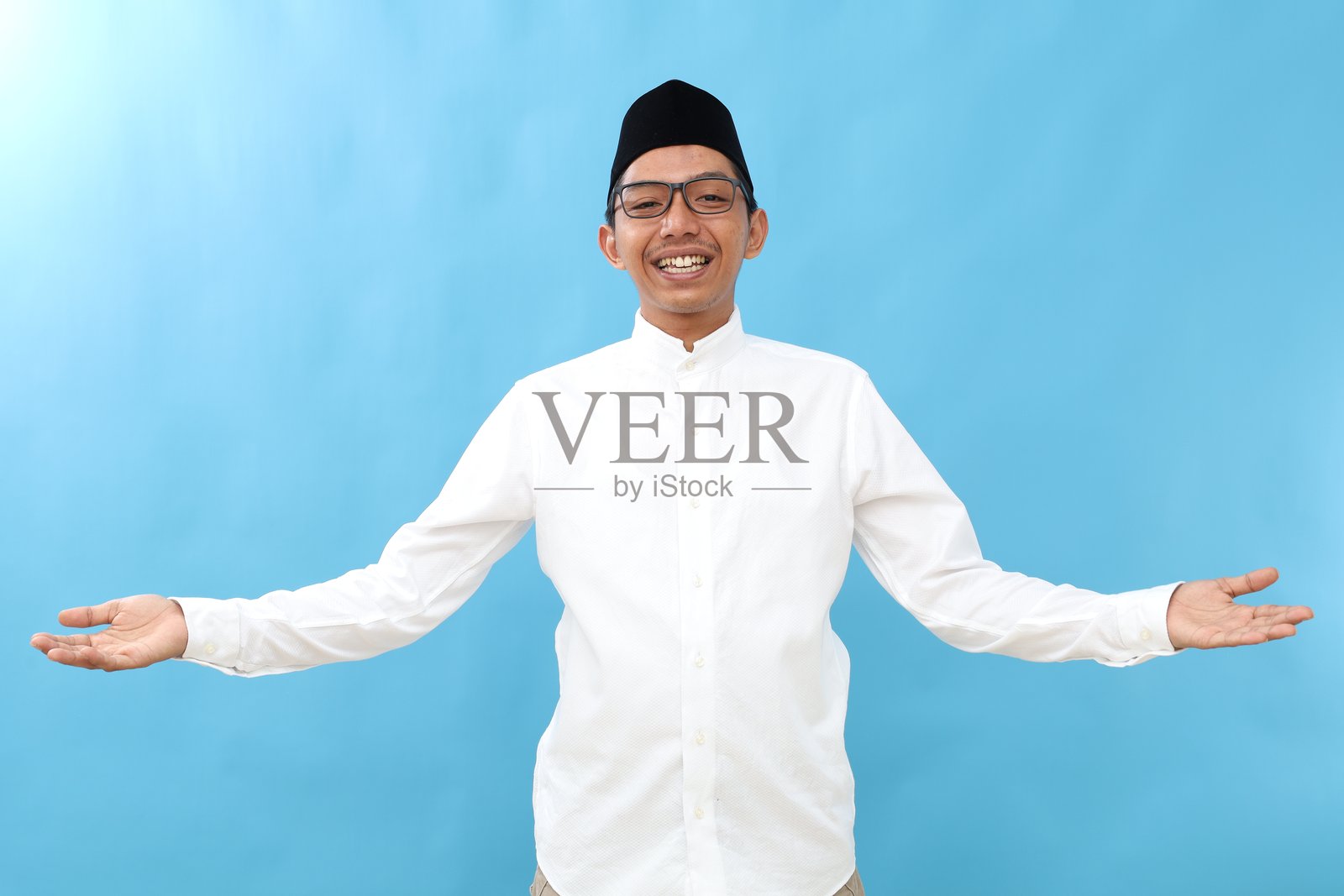 亚洲穆斯林男子在蓝色背景下微笑着张开双臂，愉快地迎接斋月庆祝活动。照片摄影图片