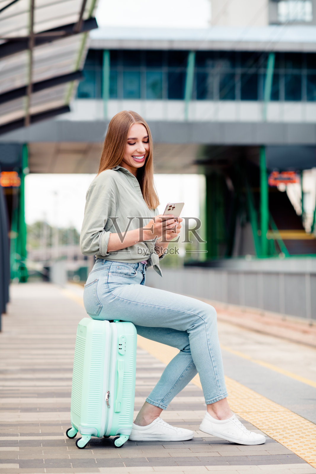 年轻女性坐在行李箱上，微笑着看着智能手机，身处城市车站的户外。照片摄影图片
