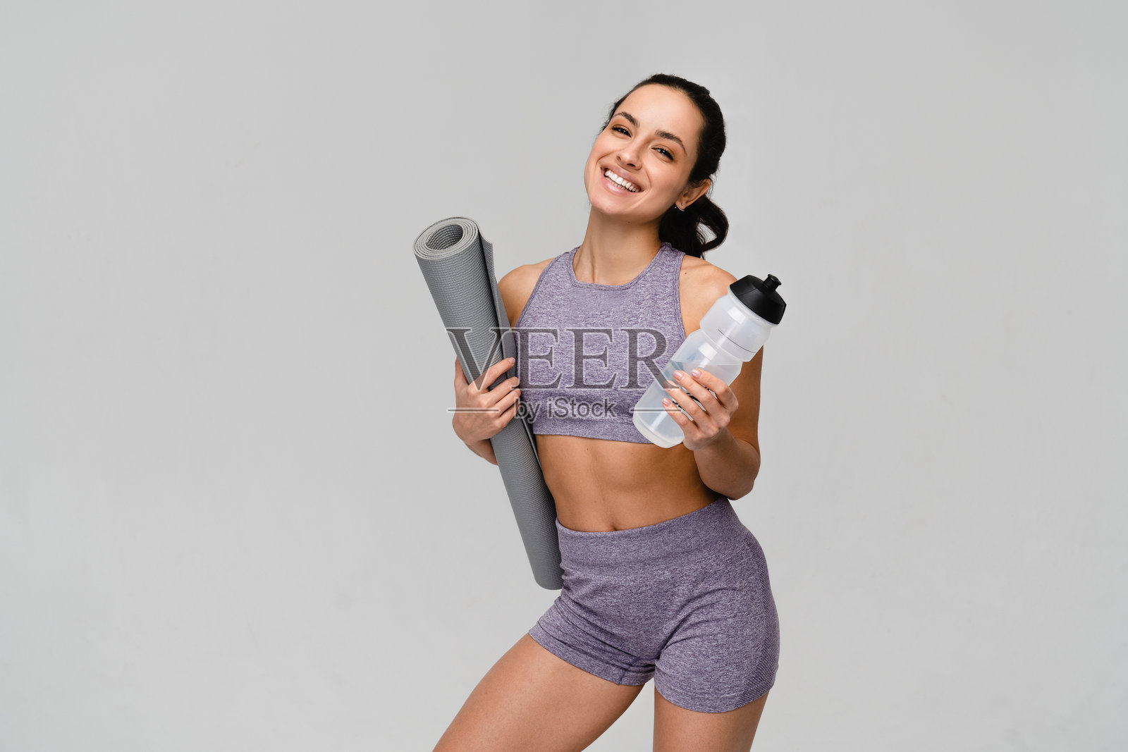 身穿运动服的女运动员健身教练，手持瑜伽垫和水瓶，背景为纯色。照片摄影图片