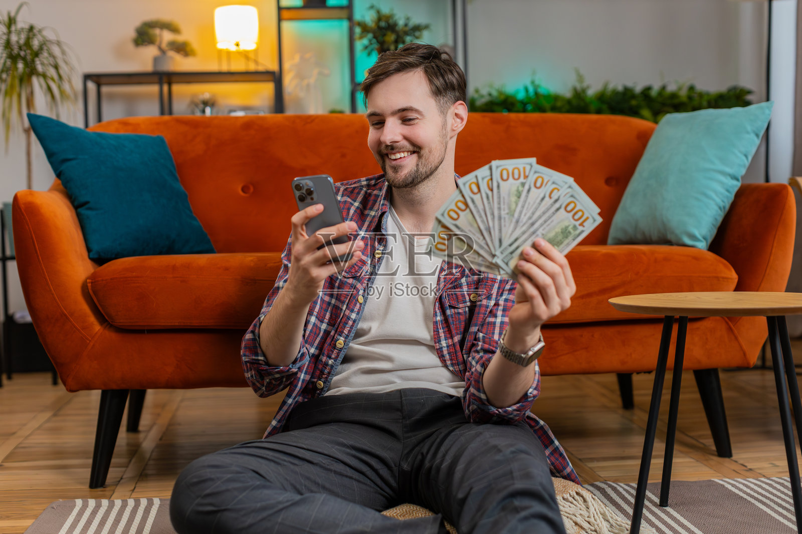 成功快乐的富人坐在家中，手持智能手机，挥舞着一叠美元钞票。照片摄影图片