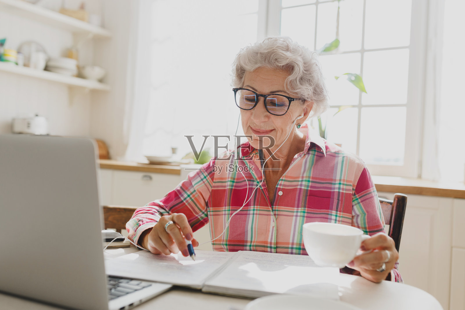 可爱的老奶奶在厨房桌子前坐着，戴着眼镜，正在在线学习，手里拿着笔，查看笔记，耳朵里插着有线耳机，手里还拿着一杯咖啡。照片摄影图片