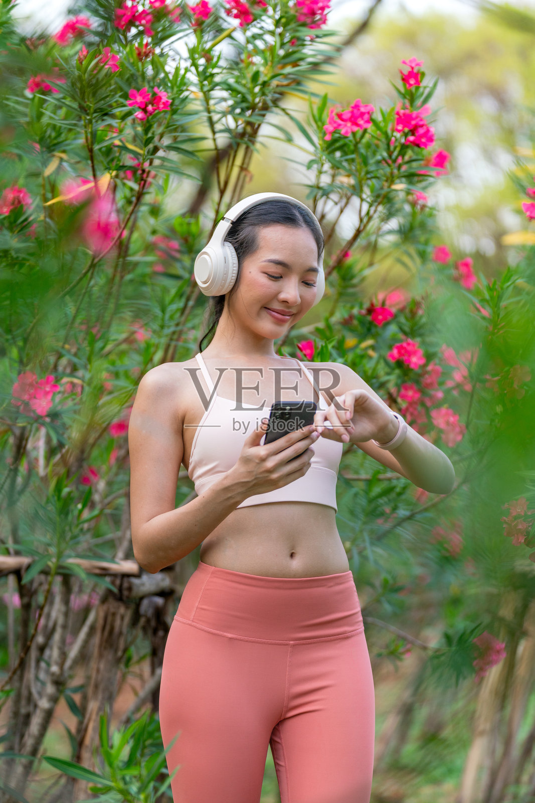 年轻亚洲女性穿着运动服，戴着耳机，在盛开的公园里锻炼时使用手机照片摄影图片