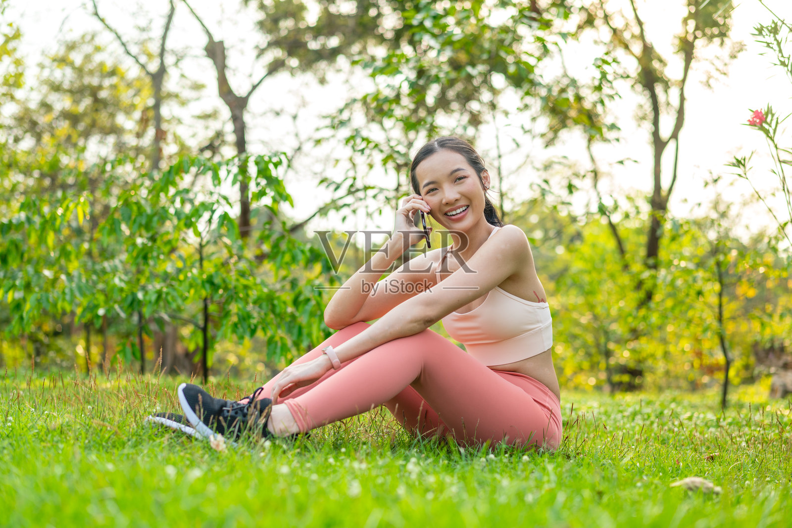 年轻的亚洲女性在公园的草地上坐着，锻炼后打电话。照片摄影图片