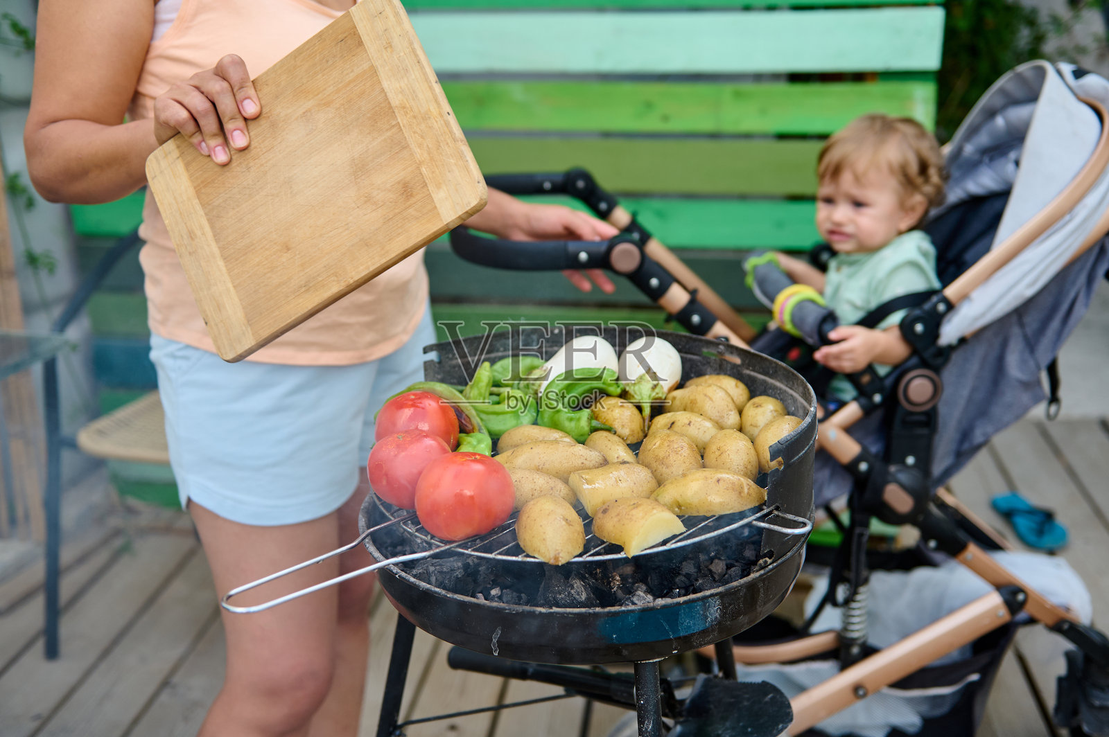 家长在户外烤蔬菜，孩子坐在旁边的婴儿车里。照片摄影图片