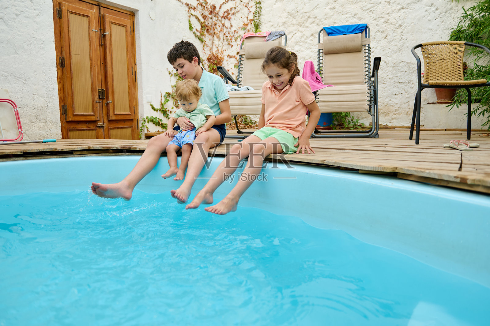 三个微笑的孩子在阳光明媚的户外泳池旁坐着照片摄影图片