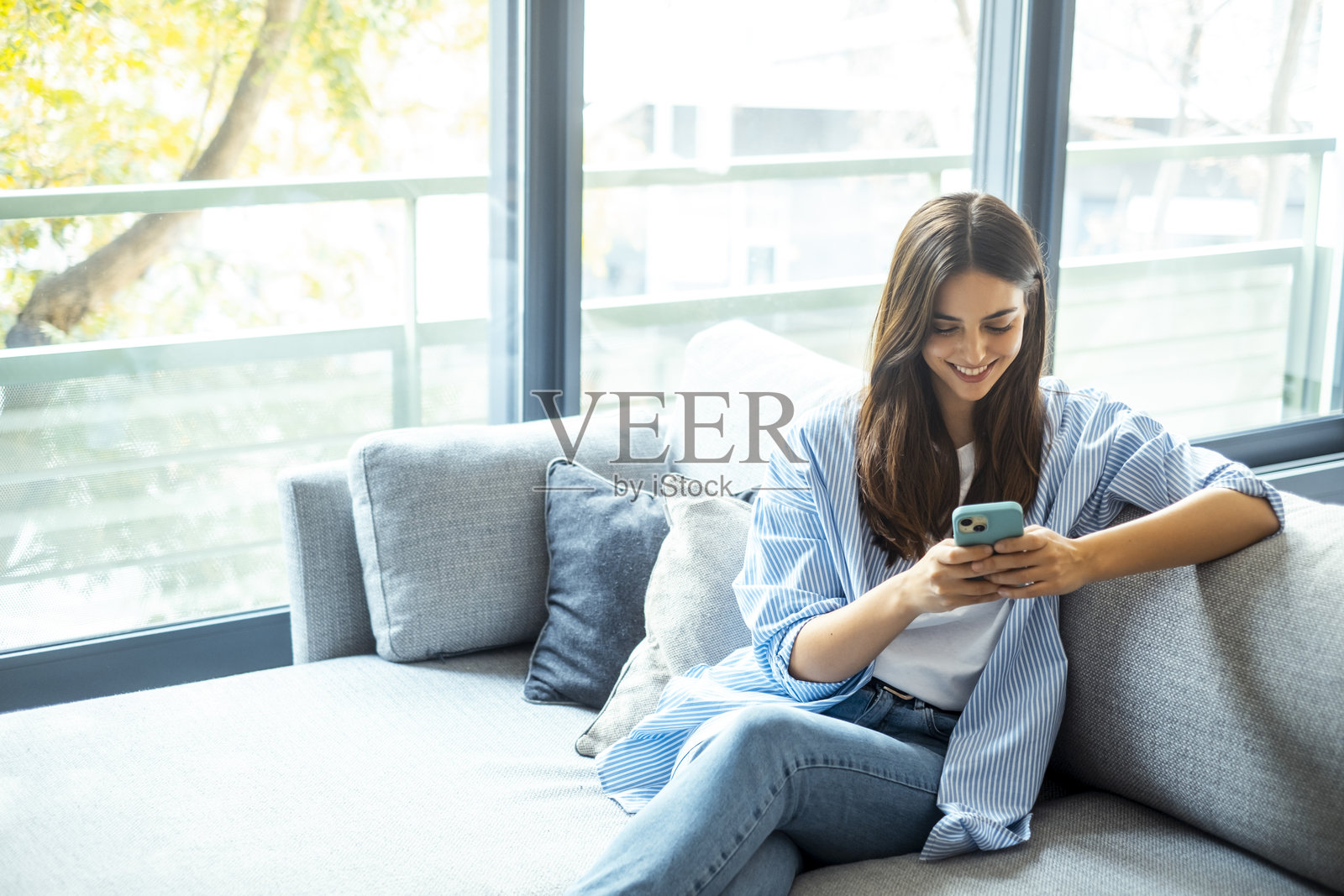 年轻女孩坐在家里的沙发上微笑，使用智能手机进行社交媒体、在线聊天、信息搜索、阅读数字内容、订阅服务和浏览互联网。照片摄影图片