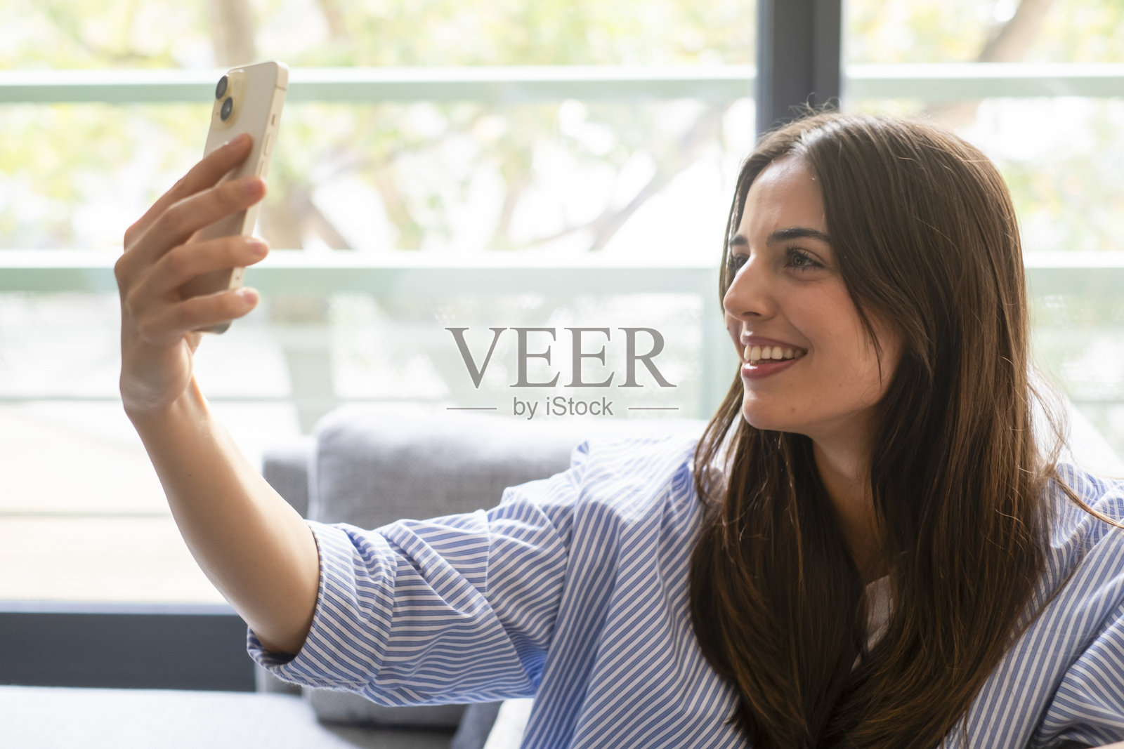 千禧一代女孩在家中坐在沙发上，放松微笑，使用智能手机浏览社交媒体、聊天应用、阅读数字内容、浏览网页、订阅服务和进行研究。照片摄影图片