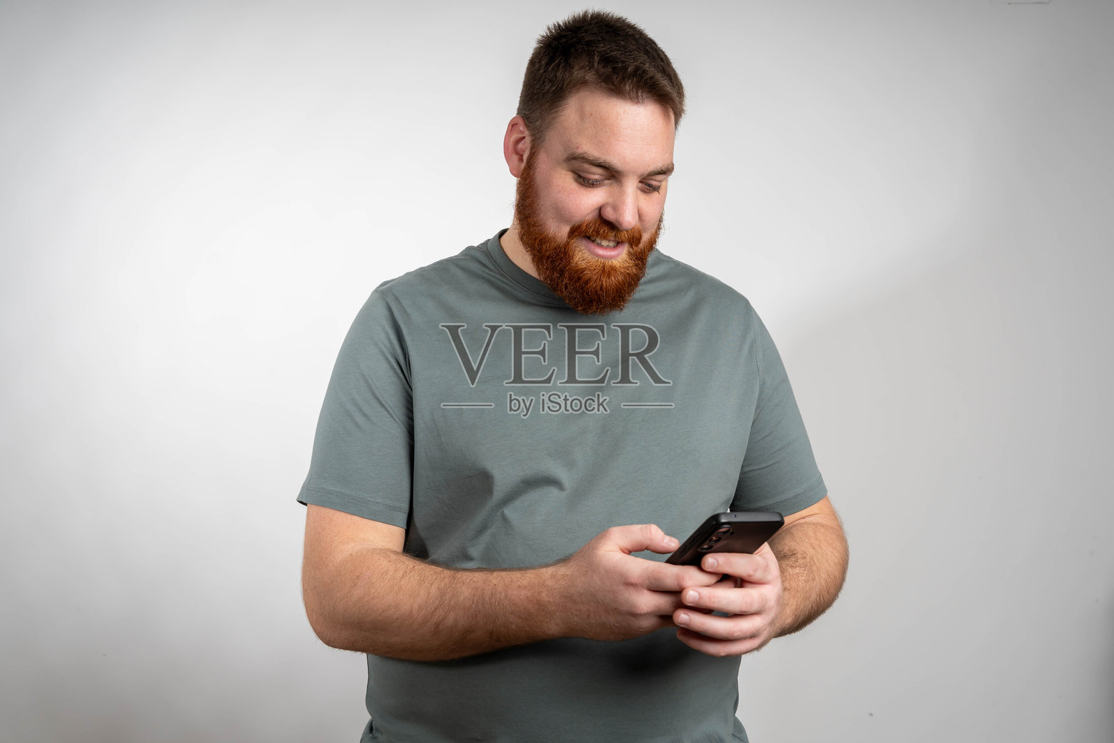 自信、积极、英俊的年轻男子，手持智能手机，在线聊天，浏览互联网，面带愉悦的微笑，站在纯白色背景前。照片摄影图片