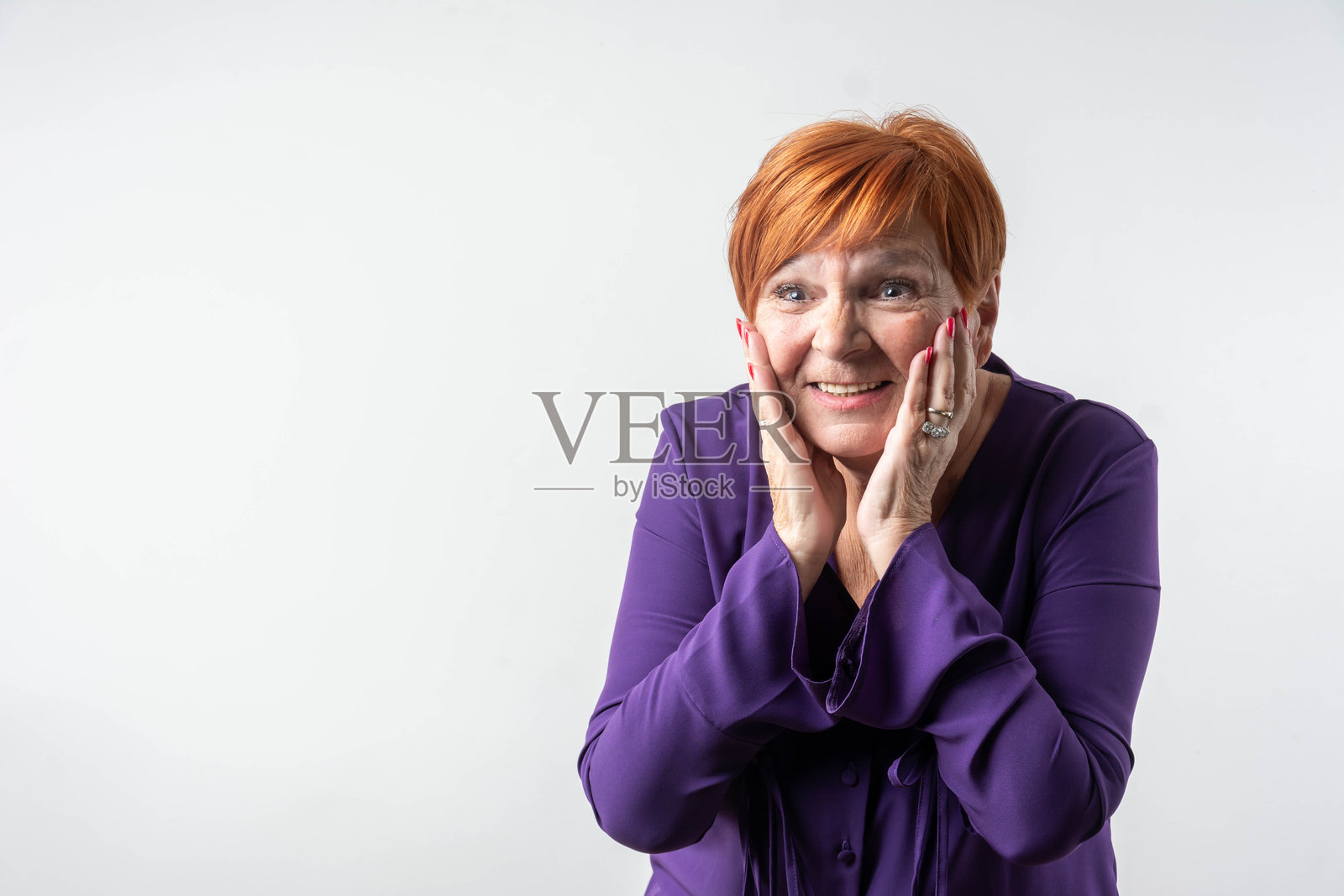 一位积极的银发女性，红色头发，穿着紫色衬衫，在灰色背景上高举双手欢庆。照片摄影图片